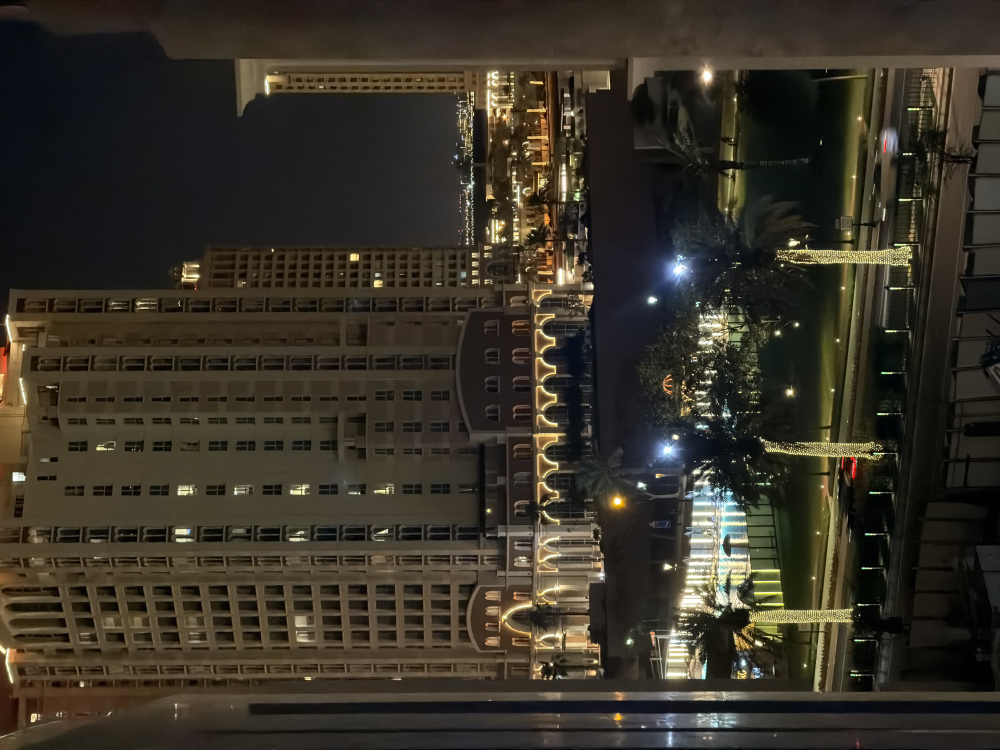 This image, captured at night from an elevated vantage point, depicts a meticulously lit urban landscape. The location is identified as **The Pearl-Qatar, Doha, Qatar**.
In the foreground, partial architectural elements, likely a window or balcony frame, obscure the far left and right edges. Below, a dark, indistinct structure is present.
The mid-ground features a multi-lane roadway where two vehicles are in motion, evidenced by light streaks and blur: a car displaying a red taillight on the left and a light-colored vehicle further right. The road is bordered by a median adorned with regularly spaced, vertically oriented green glowing lights. Adjacent to the roadway, landscaped areas feature manicured green lawns interspersed with scattered warm ground lights. Numerous palm trees are planted within these areas, with several prominent trunks densely wrapped in warm yellow string lights, creating illuminated columns.
Beyond the landscaping, a body of water, possibly a canal or lagoon, reflects ambient light and structures. A small, dark umbrella-like object is visible near the water's edge. Further back, various structures are illuminated with a mix of bright white and warm yellow lights.
The background is dominated by several tall, multi-story buildings. The most prominent structure, centrally located and light-colored (possibly cream or tan), features numerous rectangular windows. Its lower levels are distinguished by a series of large, grand arches with intricate detailing, uniformly accented with warm yellow lighting. Other similar buildings are visible further in the background, to the right and left.
The far background reveals a distant, expansive cityscape or coastline, characterized by a dense scattering of twinkling artificial lights against a dark night sky. The scene is devoid of clearly discernable individuals.