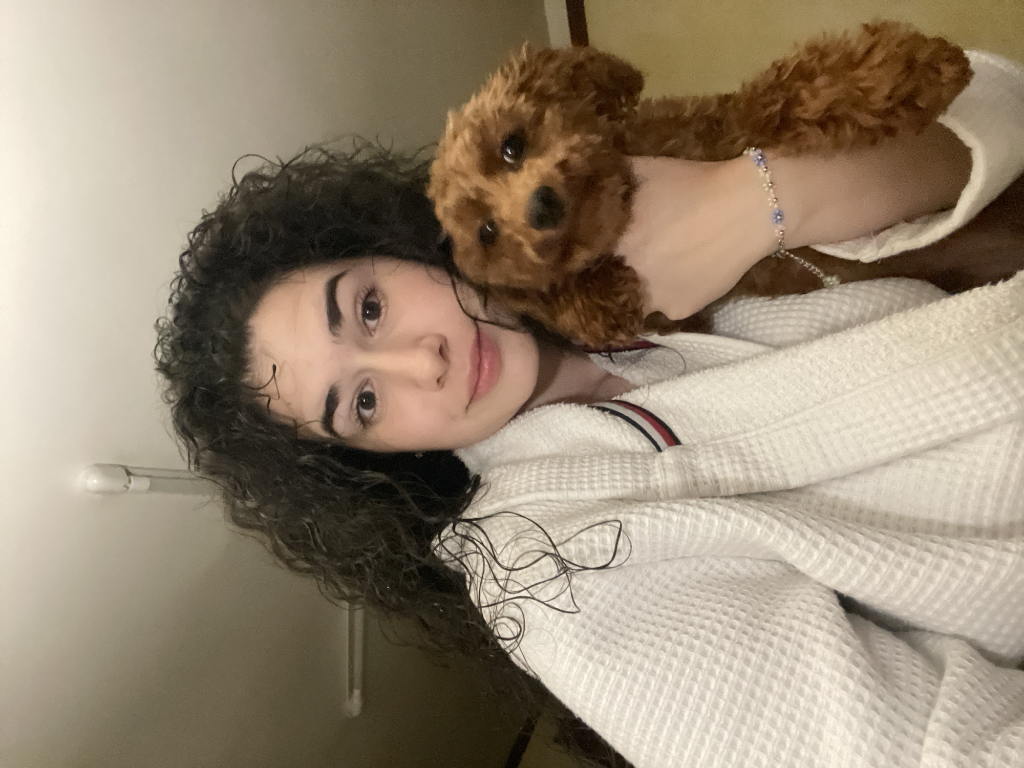 A woman with dark, curly hair is holding a small brown poodle. She is wearing a white waffle-textured bathrobe with a red, white, and blue stripe on the collar. She has a bracelet on her wrist. The background is blurry and indistinct. The location is unknown and cannot be determined from this image.