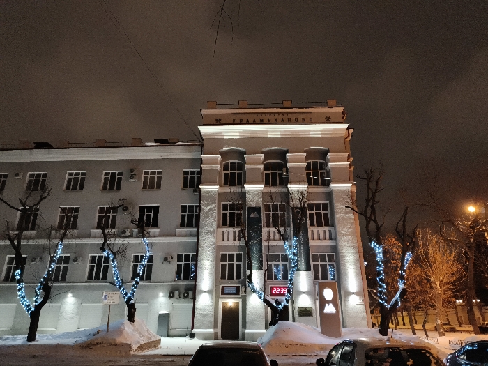A low-angle shot captures a building illuminated at night. A large, multi-story structure dominates the view, with a lighter-colored facade and prominent architectural details. Windows punctuate the exterior. bare trees are decorated with faint blue lights. Snow is visible at the base of the building, near parked cars, and a clock reads "22:33".