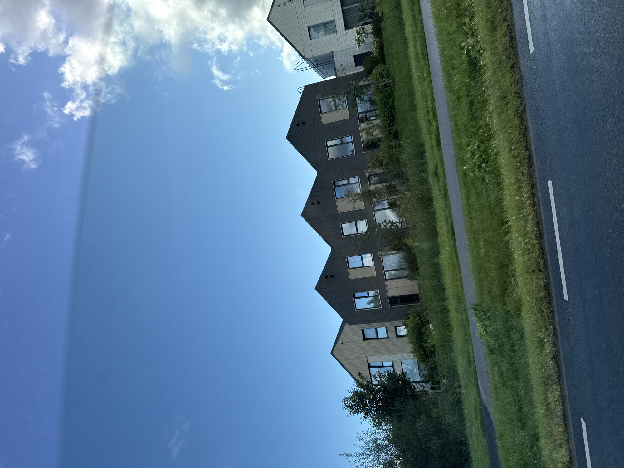 The image captures a row of modern, multi-story townhouses with dark and light facades, characterized by distinctive gabled roofs. The architecture features large windows, some reflecting the blue sky and clouds. The houses are situated on a grassy slope with mature trees and bushes in the foreground, bordering a paved pathway and a road marked with white lines. The sky is a vibrant blue with scattered white clouds. The scene appears to be a residential area in the city of Harkujärve, Estonia, likely during daylight hours. No people are visible in the frame.