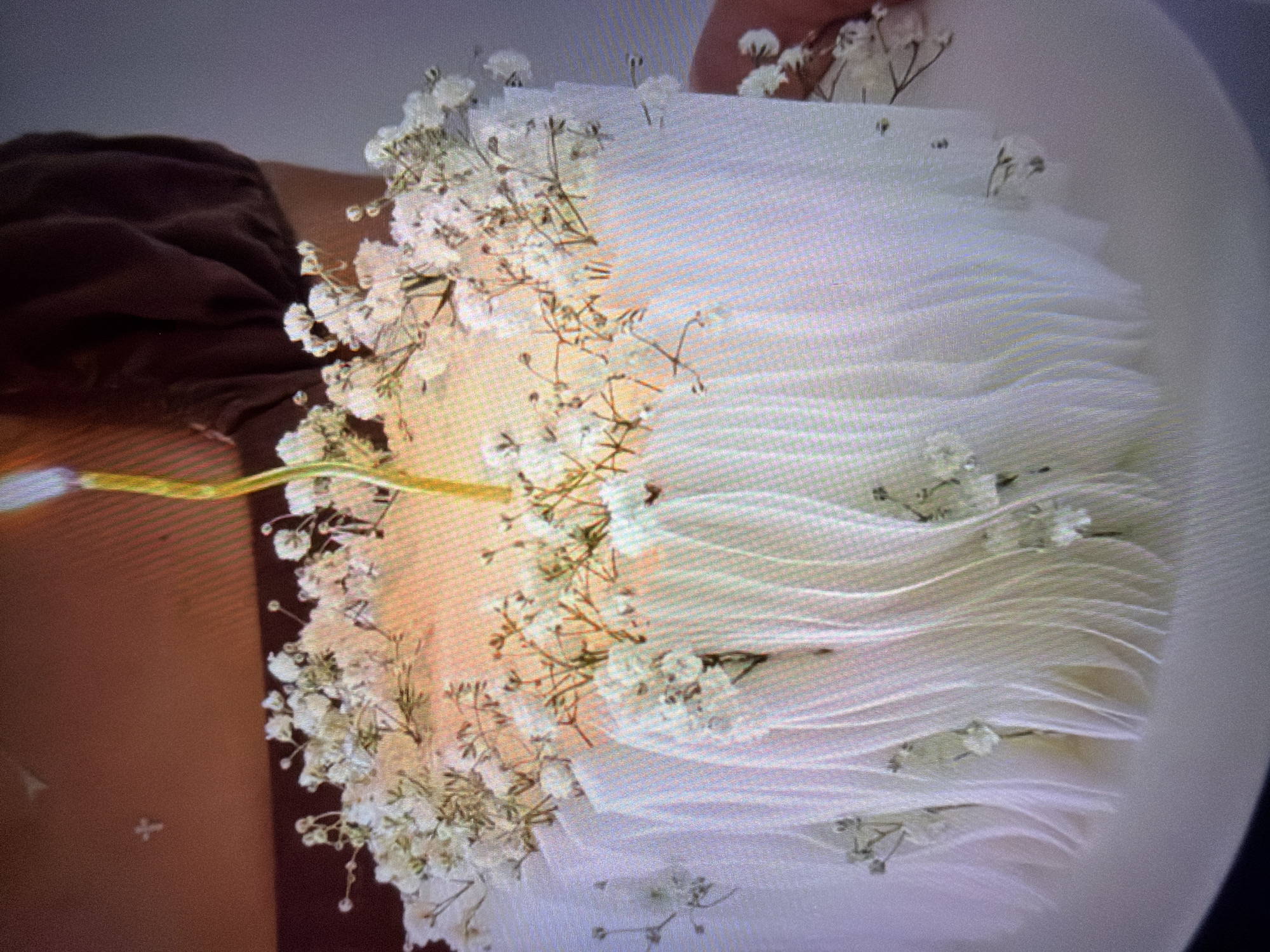 A close-up shot shows a person holding a bouquet of white baby's breath flowers and white fabric. The fabric is draped in a way that resembles layers or pleats, creating a soft, textured appearance. The baby's breath flowers are scattered throughout the fabric, with some clusters more dense than others. The stem of the bouquet is visible, appearing to be made of a yellow material. To the left of the bouquet, a person's arm is visible, covered by a dark brown garment, suggesting a warm and possibly casual setting. The background is a mix of soft, muted tones, with a hint of a light source casting a warm glow on the fabric and flowers. The overall impression is one of delicate beauty and simplicity, possibly for a wedding or special occasion. The image is somewhat blurry and pixelated, suggesting it might be a photograph taken from a screen or a lower-resolution digital capture. There is no visible text in the image.