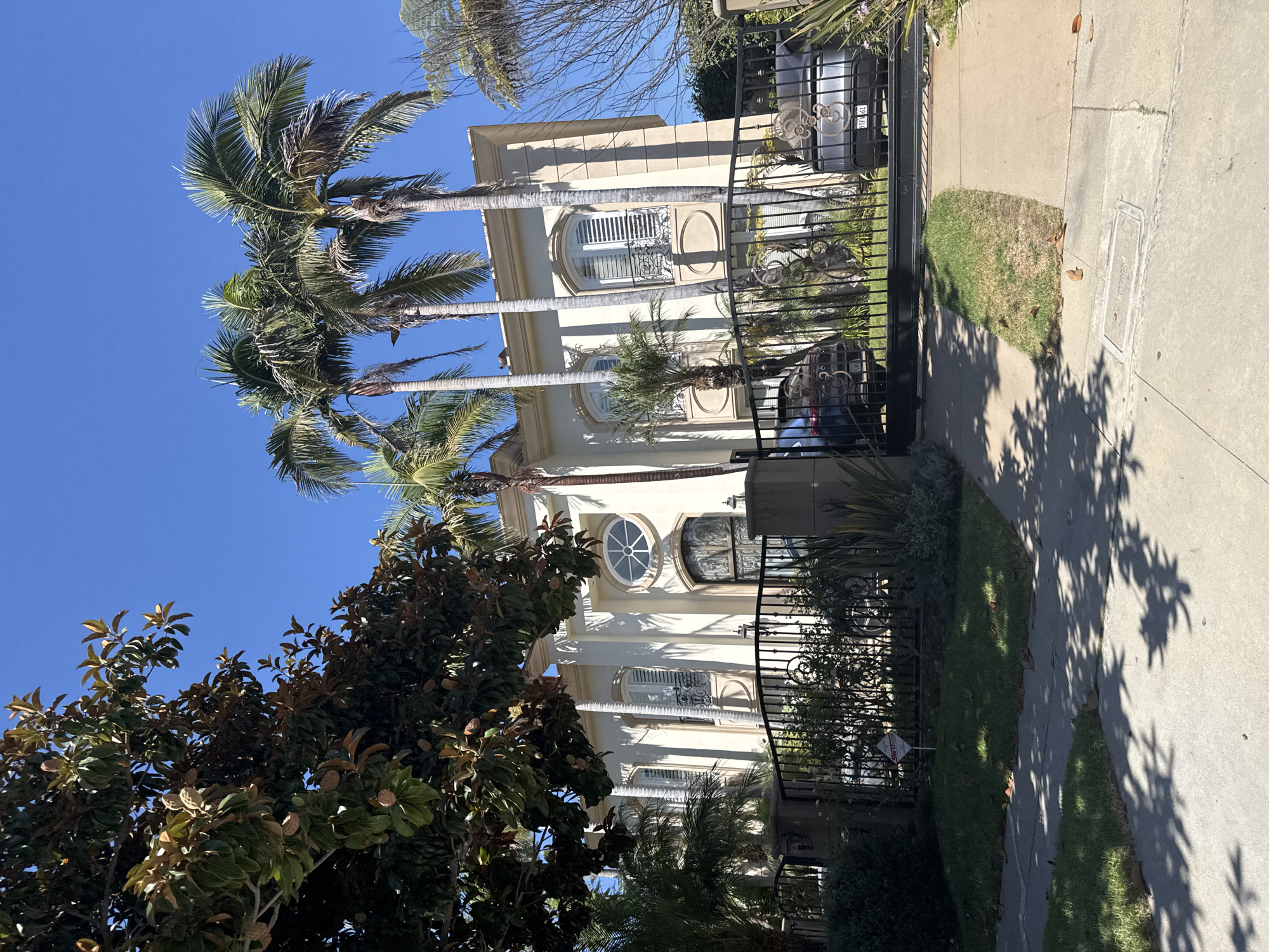 The image depicts a large, ornate mansion in Beverly Hills, California, United States of America. The house is painted a light cream color and features classical architectural elements such as columns, arched windows with shutters, and decorative ironwork. Several tall palm trees and a mature magnolia tree with large seed pods are visible in the front yard, casting shadows on the property. A black wrought-iron gate, adorned with decorative scrolls, stands in front of the driveway, which has two cars parked behind it. A red and white "SWAT" security sign is visible on the lawn in front of the gate. The sky is clear and blue, indicating a sunny day. The sidewalk and a small patch of grass are in the foreground.