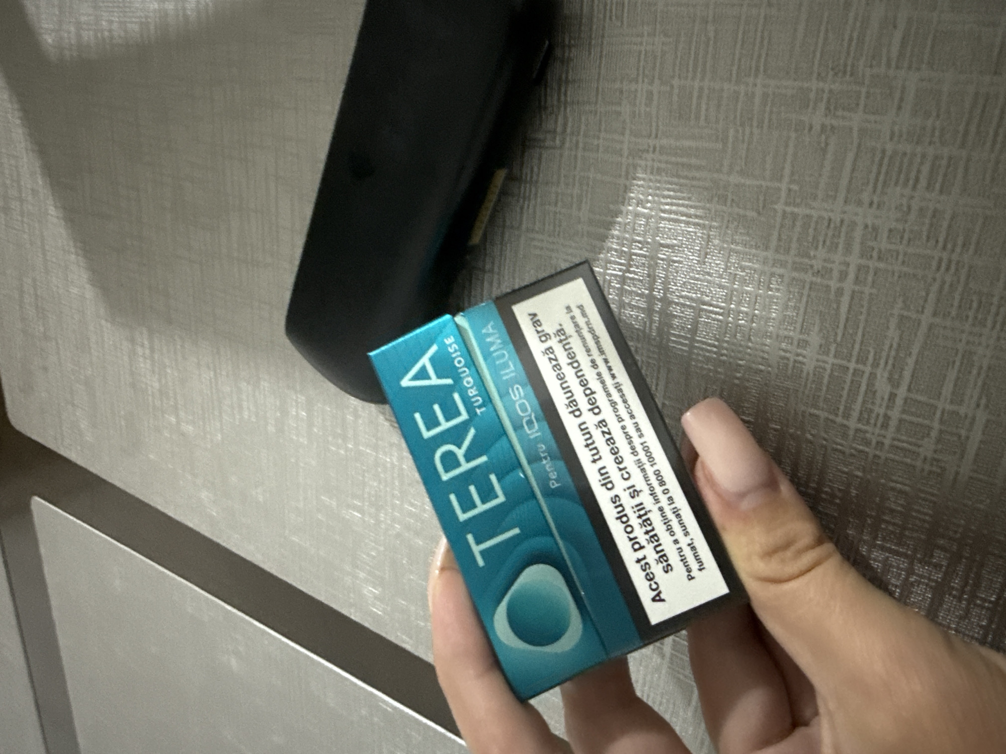 The image shows a close-up view of a hand holding a pack of Terea Turquoise IQOS ILUMA tobacco sticks. The pack is teal and white with text in Romanian. The text translates to "This product is harmful to health and causes addiction. To obtain information about the programs to quit smoking, call 0 800 10001 or visit www.imspsm.md". In the background, a black IQOS device is visible on a textured surface that appears to be a table. The lighting suggests an indoor setting. The context of the image does not provide any specific information about the location in Chișinău, Moldova.