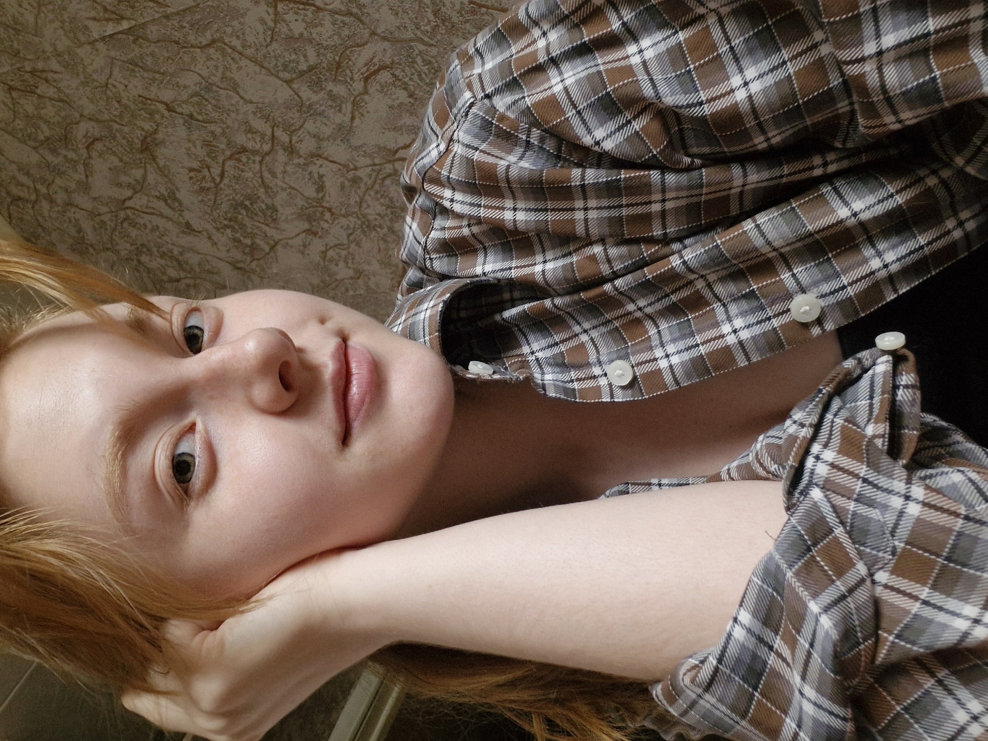 The image is a close-up shot of a woman with red hair, fair skin, and light-colored eyes, lying on her side with her head resting on her hand. She is wearing a buttoned-up plaid shirt in shades of brown, gray, and white. The background appears to be a textured wall with a patterned surface. The overall impression is a casual, relaxed pose. There is no evidence to suggest the image was taken in Yekaterinburg, Russia, or any other specific location.