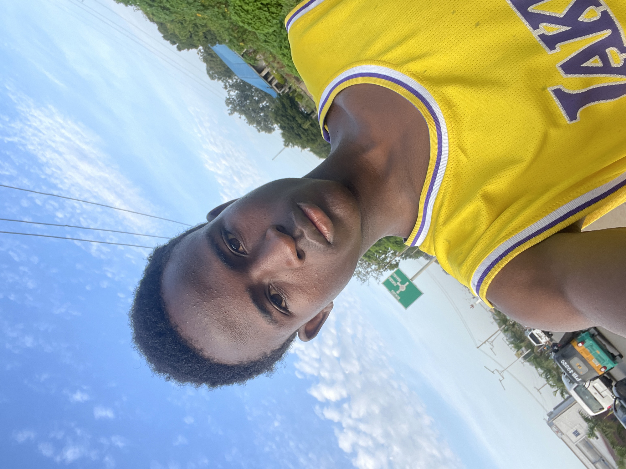 In a selfie taken outdoors, a young Black male, appearing to be in his late teens or early twenties, is pictured against a backdrop of a bright, partly cloudy sky. He is wearing a yellow basketball jersey with purple and white trim, and "LAKERS" is visible in large purple lettering across the chest. His hair is short and black, and he has dark eyes and brown skin. The background reveals a busy street scene with a green road sign indicating "BUKURU" and "DANGOGO." Various vehicles, including a white van, a teal and yellow auto-rickshaw with "PTA-SOURO" written on it, and other cars, are visible on the road. Trees and some buildings with blue roofs are also present, suggesting an urban or semi-urban environment. The lighting indicates it is daytime. This scene is consistent with an outdoor location in Jos, Nigeria, given the presence of "Bukuru" and "Dangogo," which are areas within or near Jos.