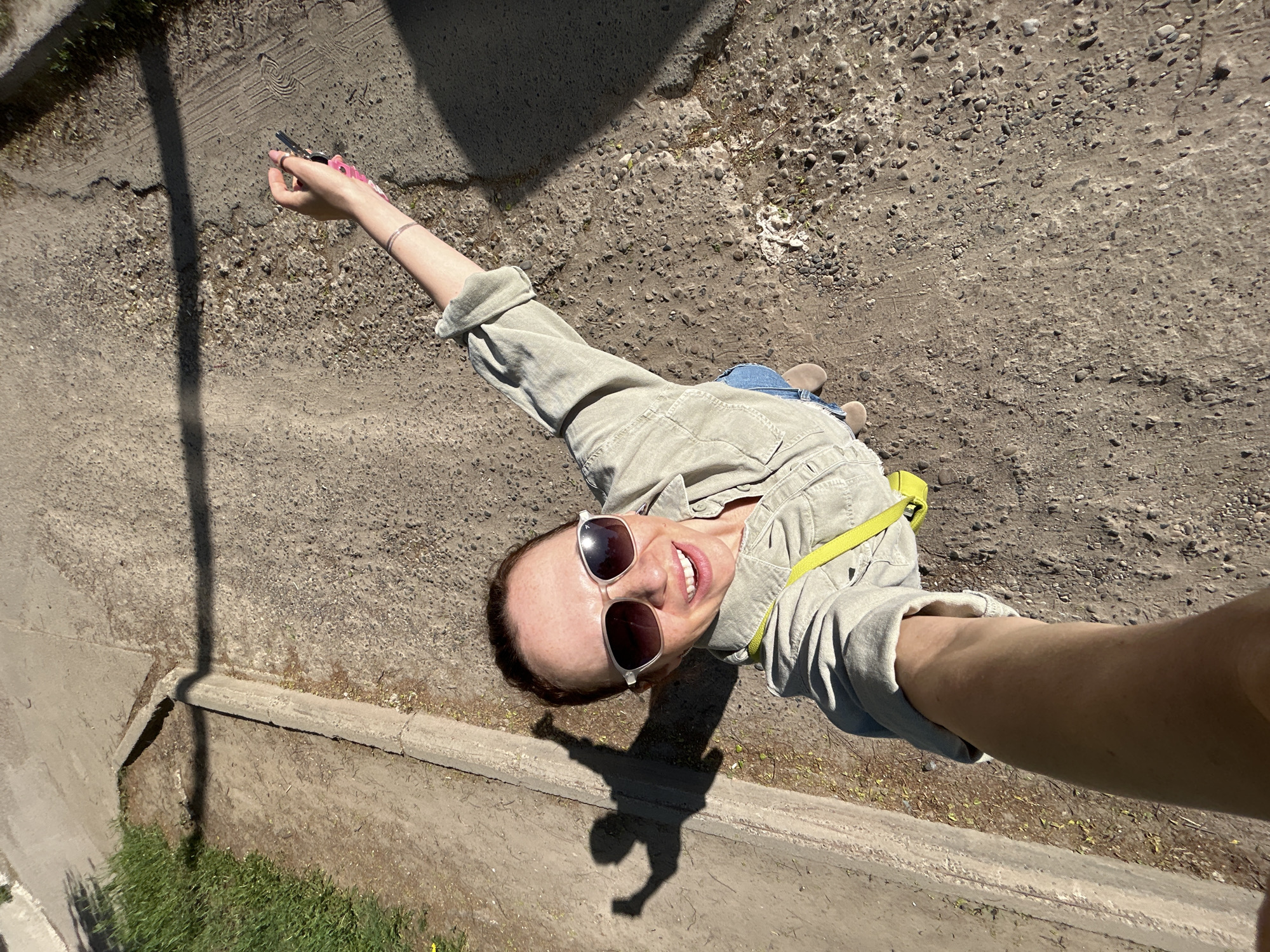 A high-angle perspective shows a woman taking a selfie outdoors on a sunny day. She is facing the camera and smiling, wearing sunglasses and a light-colored button-up shirt, with a yellow strap visible across her shoulder. Her right arm is extended toward the camera holding a phone. In her left hand, she holds a set of keys with pink accessories. The ground appears to be a paved or unpaved area with a mix of concrete, gravel, and dirt. The woman's shadow and a possible lamppost shadow are also visible on the ground. Part of a low concrete structure with grass growing along it is visible along the left side of the frame. The location is the city of Chaikovsky, Russia.