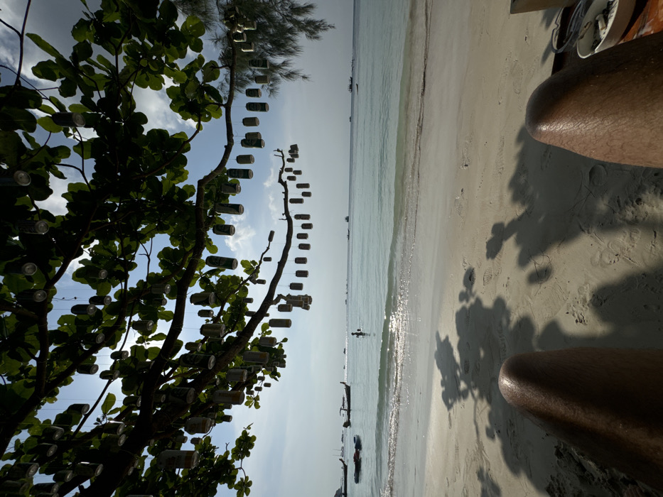 The image depicts a first-person perspective of a tropical beach scene in Unknown, Unknown. In the immediate foreground, a person's bare, tanned legs are visible, extending into the lower portion of the frame and resting on light-colored sand. Tree foliage casts dark shadows across the sand, which also shows faint footprints. A white ashtray containing cigarette butts is partially visible on the far lower right.
The sandy beach transitions into calm, clear turquoise ocean water with small breaking waves. One individual is partially submerged, standing in the water in the mid-distance. To the left, two brightly colored floating objects, resembling kayaks or paddleboards, are present near the shore. Several traditional long-tail boats are anchored or moving in the mid-distance on the water. The horizon line separates the ocean from a bright sky with scattered white clouds.
Overhead, dense green foliage from a tree extends into the upper frame. Numerous empty aluminum beverage cans, predominantly green and silver, are suspended by lines from these tree branches, serving as decorative elements. The scene is illuminated by strong sunlight, creating a slight backlight effect through the foliage.