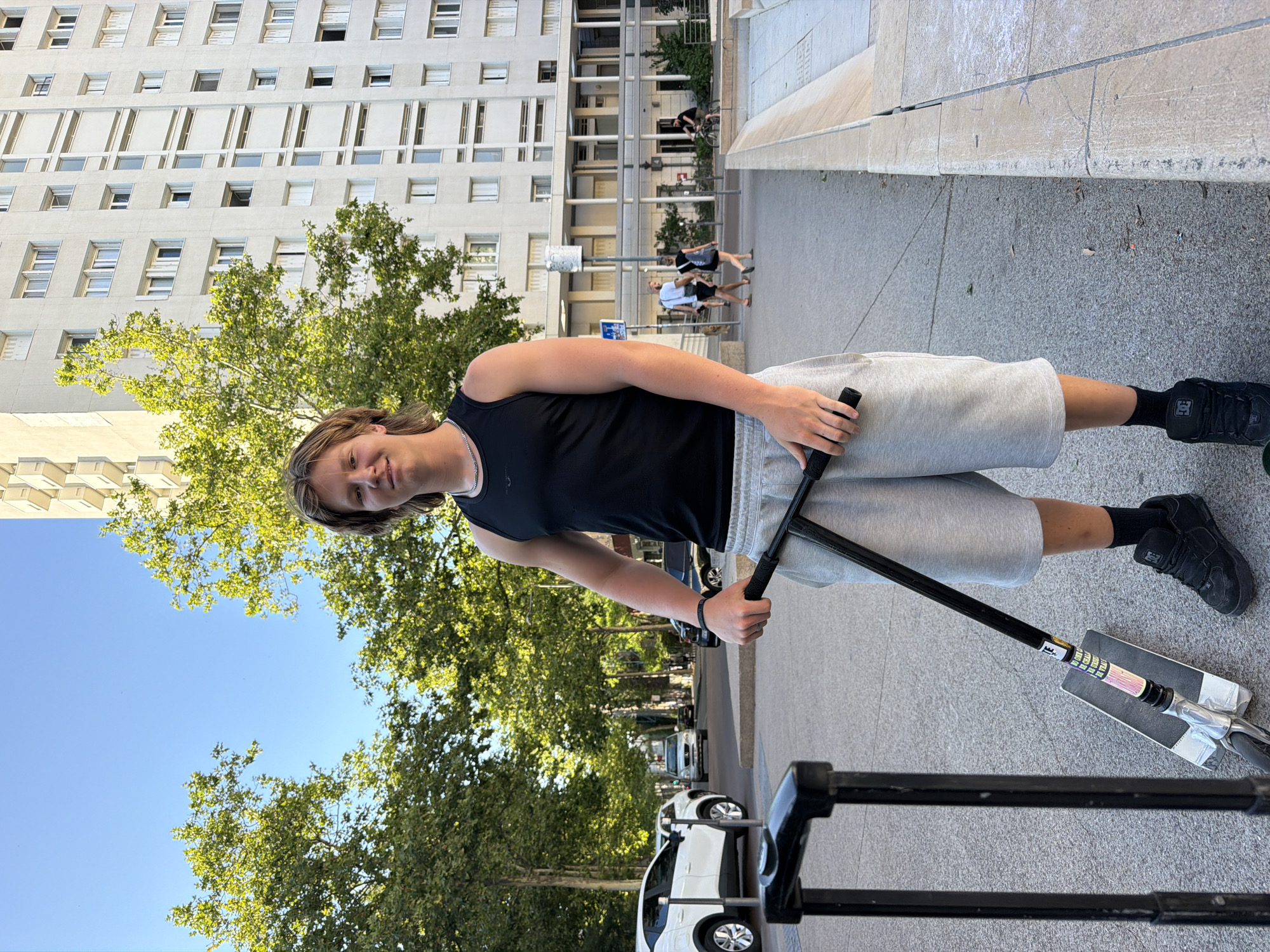 A young male with blond hair, wearing a black tank top and grey shorts, stands on a paved surface in front of a tall, white apartment building. He is holding the handlebars of a scooter. To his left is a black suitcase. In the background, trees line a street with cars and pedestrians. A man on a bicycle is visible further down the street. The building in the background appears to be part of the "Les Géraniums" residence located in the Vaulx-en-Velin district of Lyon, France. The overall scene depicts a sunny urban environment.