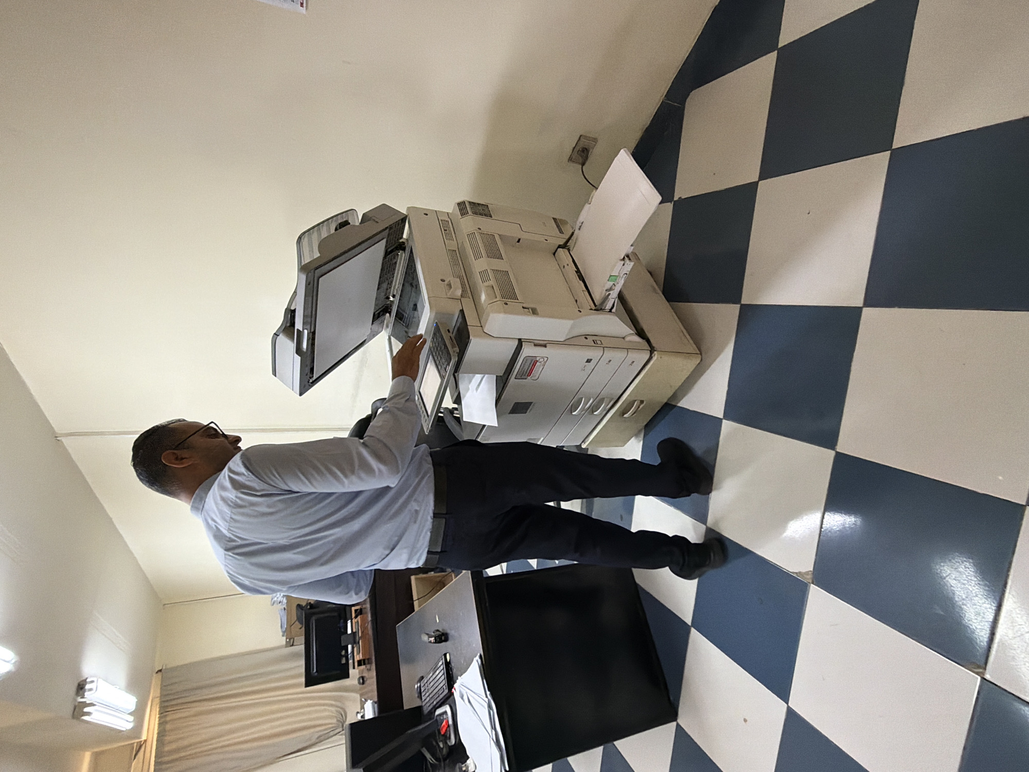 In a dimly lit office space in Sharm El Sheikh, Egypt, a man wearing a light blue collared shirt and dark trousers stands in front of a large, beige and white office copier. The copier's lid is open, and the man's hands are near the control panel and the paper feed tray. He appears to be operating or loading the machine. The office floor is tiled in a checkerboard pattern of dark blue and white. To the left of the copier, a dark desk with a computer monitor and keyboard is visible. In the background, cream-colored walls are adorned with ceiling-mounted fluorescent lights and sheer curtains. The scene suggests a typical office environment where document processing is taking place.