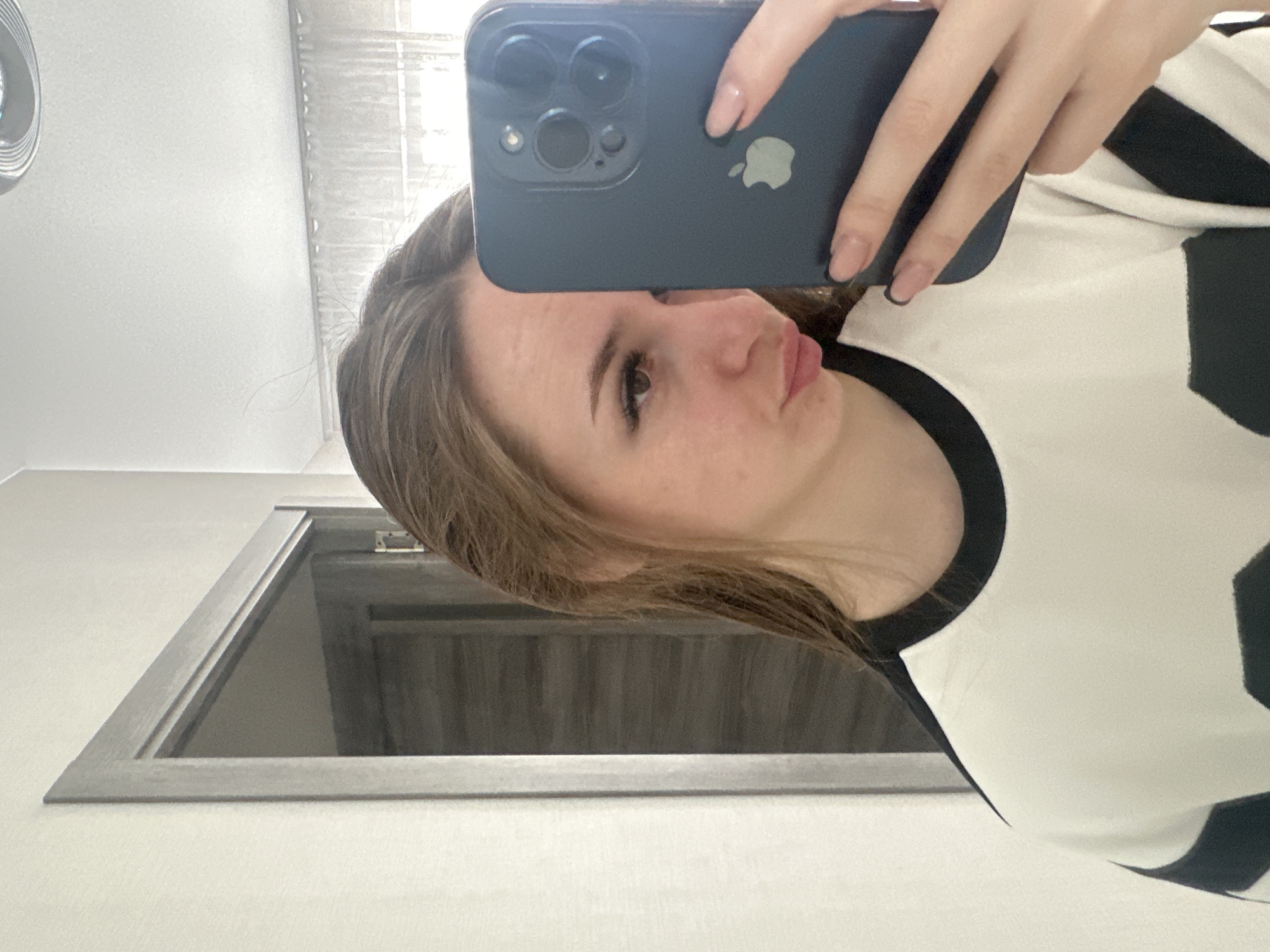 A young woman is captured in a mirror selfie, holding a dark-colored smartphone to take the picture. The image is oriented sideways, with the woman's head tilted. She has light brown hair, and only the right side of her face is clearly visible, showing her eye, nose, and lips, which are slightly puckered. She wears a white top with bold black stripes or patterns, and a black trim around the neckline. Her right hand holds the iPhone, revealing a prominent triple camera system and a silver Apple logo on its back. Her nails are manicured with a light, natural-looking polish.
The background suggests an indoor setting with light-colored walls and a white ceiling. A silver, circular light fixture is partially visible in the top left corner (when the image is rotated upright). Behind the woman, a dark-framed doorway or opening is present, revealing what appears to be vertical blinds or a textured wall in the adjacent space. The overall lighting suggests it is daytime.