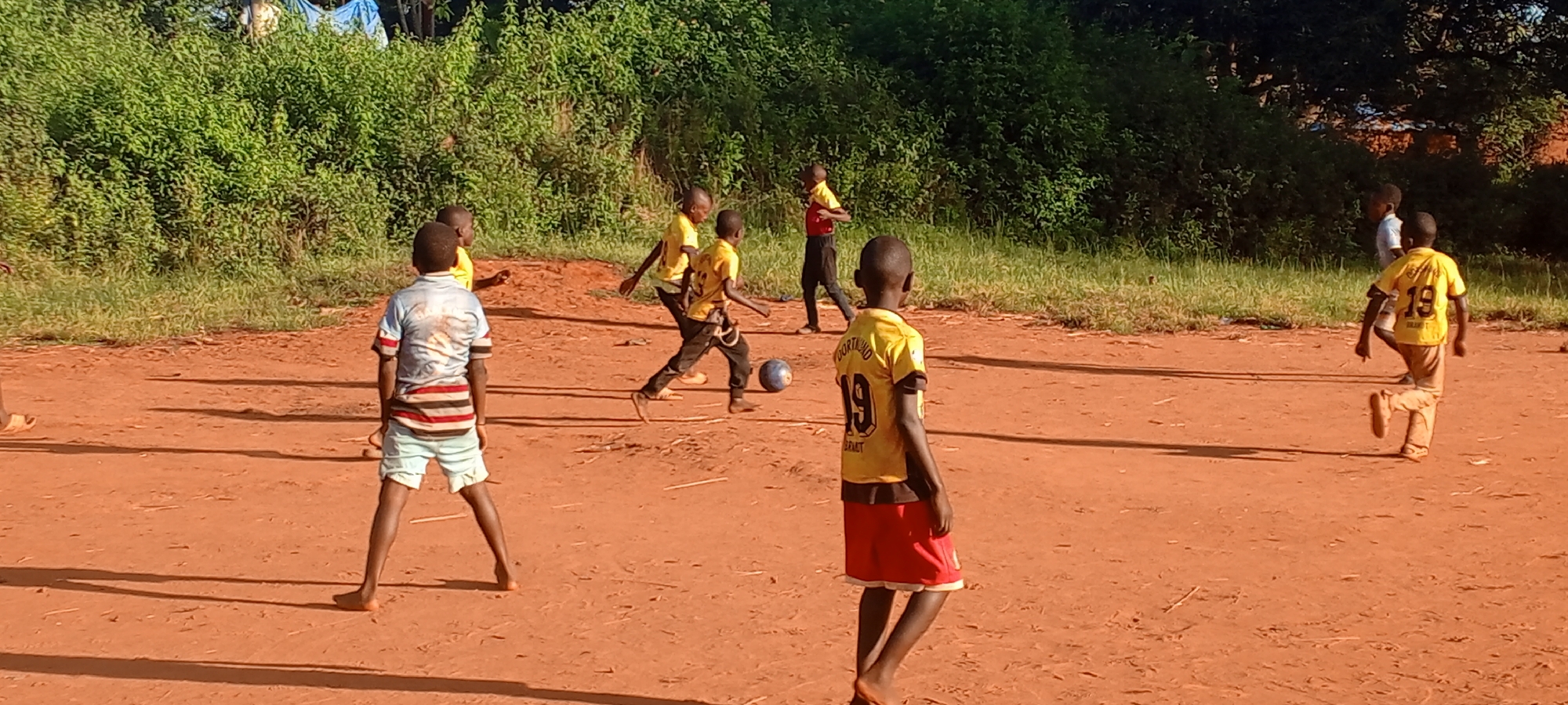 The image shows a group of barefoot children playing soccer on a dirt field in Ngalula, Democratic Republic of the Congo. The field is surrounded by lush green vegetation. Most of the children are wearing yellow jerseys with the number "19" on the back. One child is wearing a red and yellow jersey, while another is wearing a blue striped shirt and light-colored shorts. One child is actively kicking the soccer ball. Several other children are watching the game.