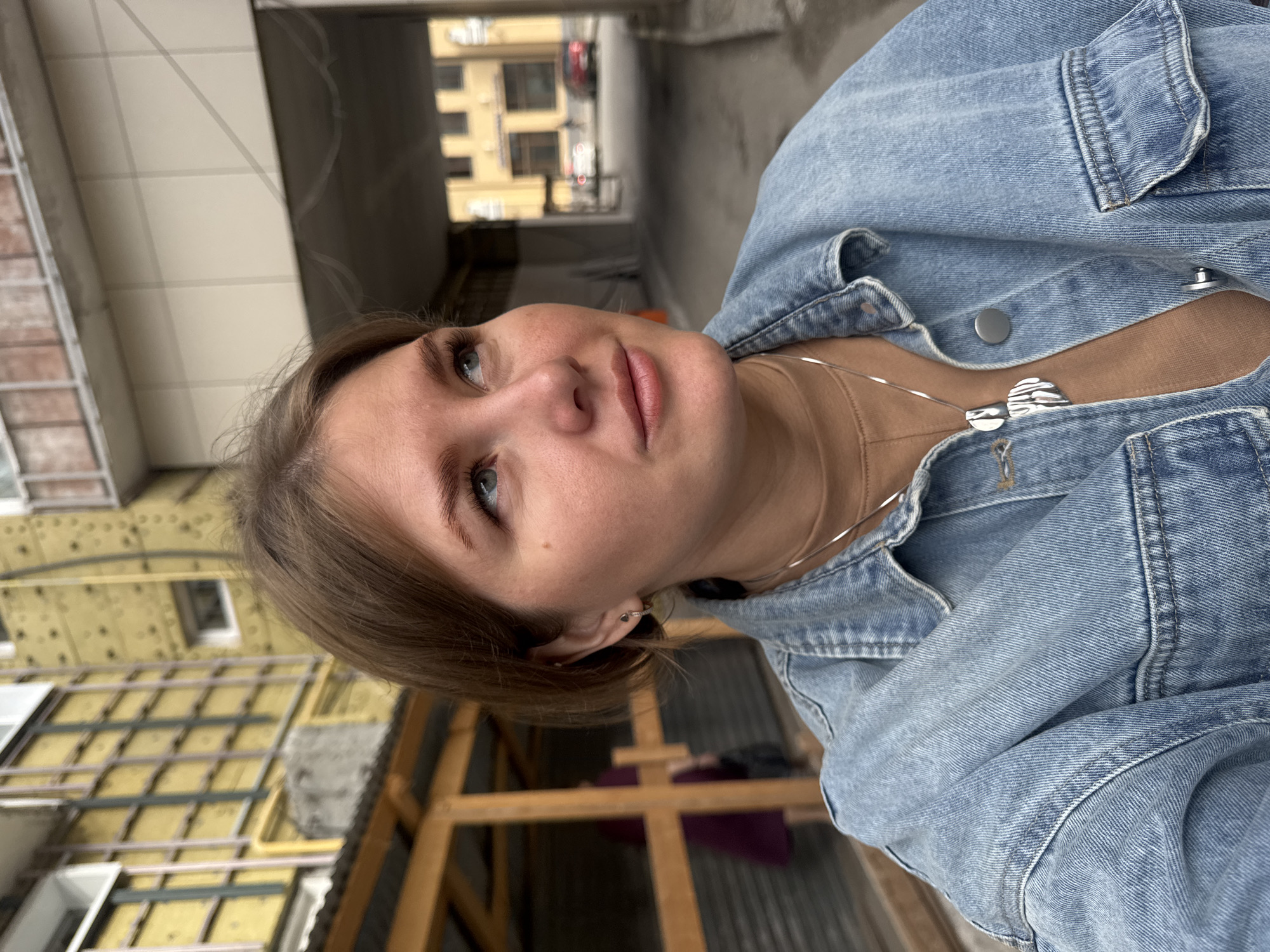 A fair-skinned female with light brown hair is pictured wearing a denim jacket and a silver necklace with a pendant. She has blue eyes and is looking upwards with a slightly pensive expression. The background includes a building under construction with exposed insulation and wooden framing, as well as another building in the distance. The setting appears to be an urban area, specifically in Perm, Russia.