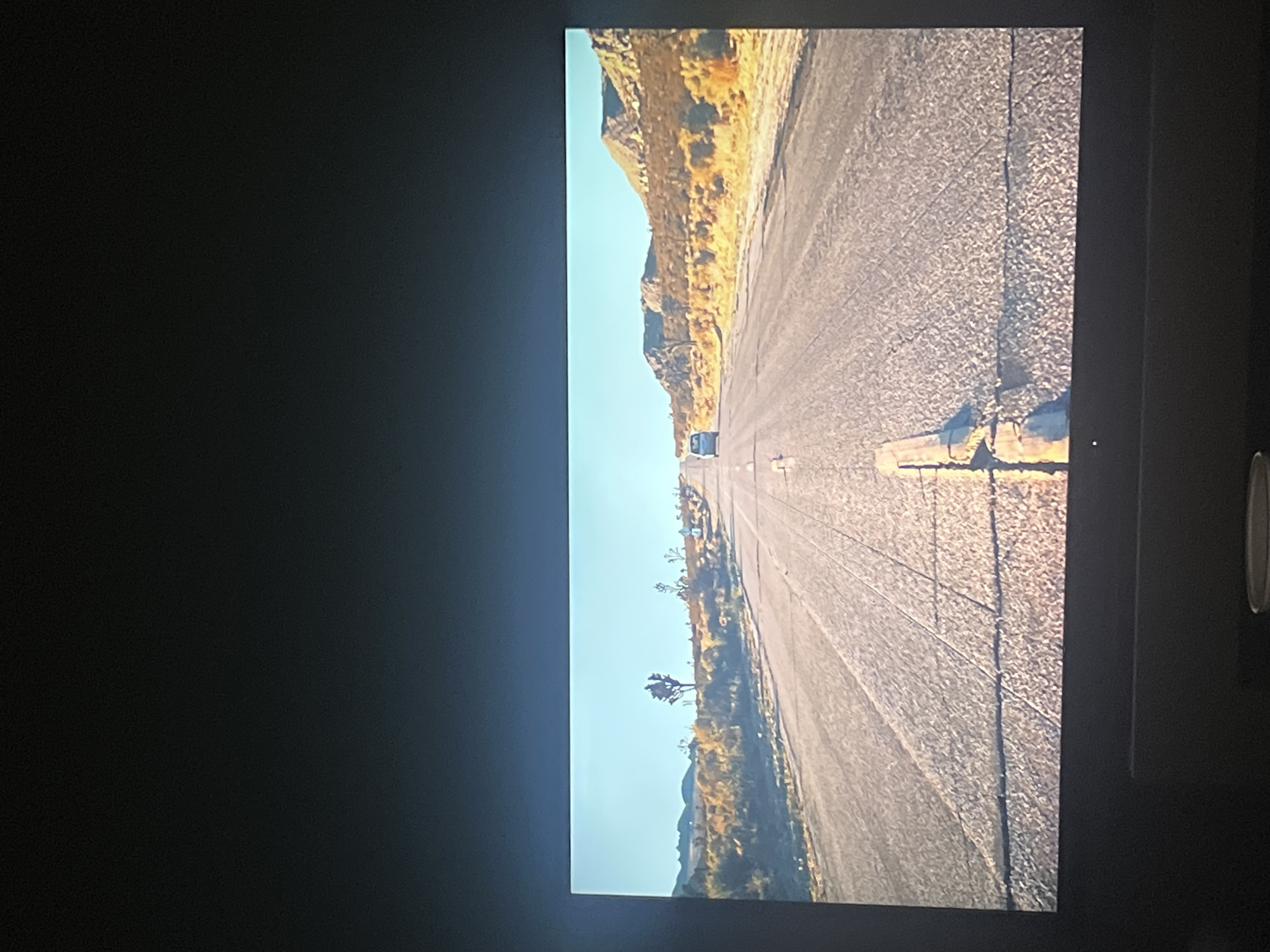 The image depicts a desert road with a vehicle in the distance. The road stretches into the horizon, bordered by desert terrain on both sides. The sky is visible above the landscape. The image is displayed on a screen within a dark room. There is no information to suggest the location is in Mexico City, Mexico.