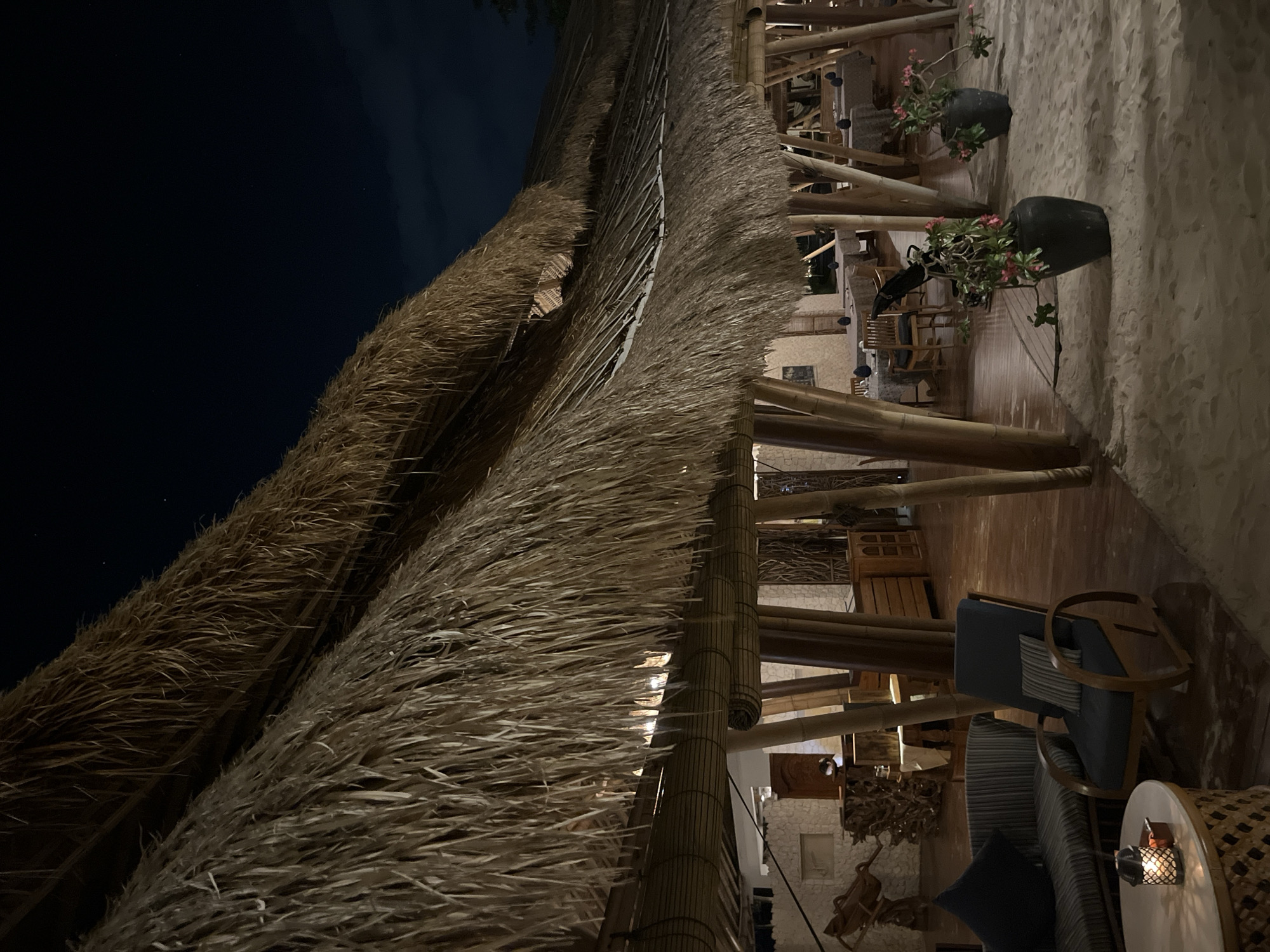 The image depicts a nighttime scene at an outdoor restaurant or resort with a thatched roof. The scene is illuminated by warm, artificial lighting, suggesting it is after sunset. The architecture features prominent bamboo structural elements and a thatched roof made of dried palm fronds, creating a rustic and tropical ambiance.
The foreground is dominated by the texture of the thatched roof, with individual fronds clearly visible. Beyond the roof, the restaurant's interior is visible, with wooden floors, more bamboo pillars, and various seating arrangements. Several tables and chairs, some made of wood and others upholstered in dark blue fabric, are arranged on the sand-covered ground and wooden platforms.
Two large, dark, urn-like pots containing flowering plants with pink blossoms are placed on the sand. A small, lit lantern sits on a round side table next to a couch, casting a warm glow and adding to the cozy atmosphere. The overall impression is one of a tranquil and inviting evening setting.
No people are visible in the image. The sky in the background is dark, indicating nighttime, and a few faint stars might be visible, suggesting clear weather. The sand and tropical vegetation, combined with the bamboo and thatch architecture, strongly suggest a location in a warm, coastal region, consistent with the provided location context of Kuta Selatan, Indonesia. The overall mood is peaceful and relaxing.