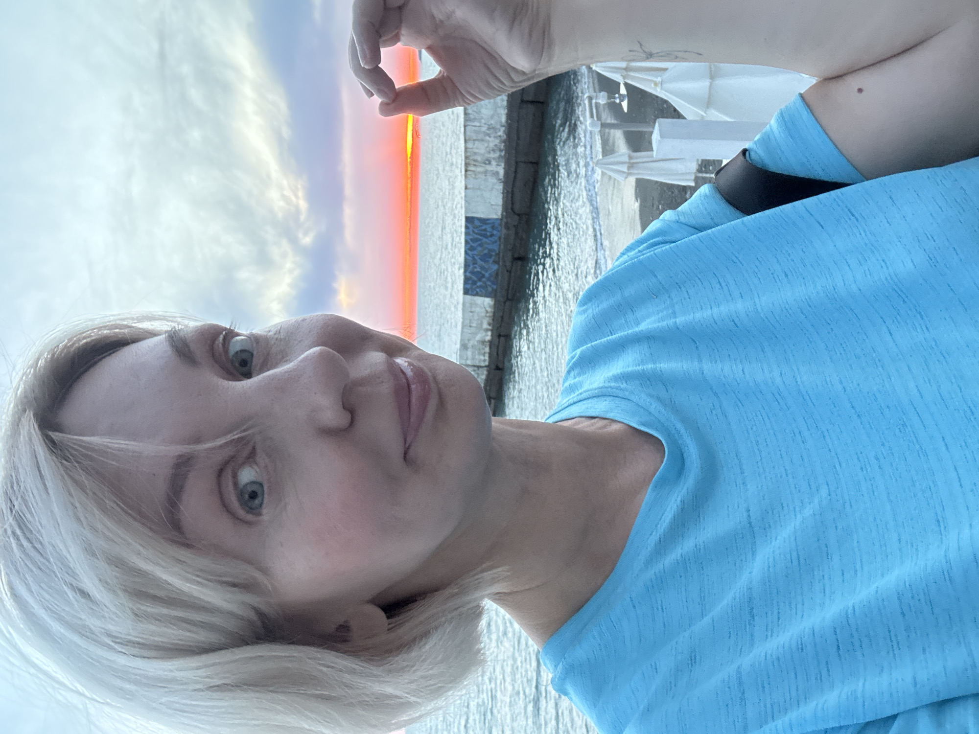 The image is a selfie taken outdoors at sunset near the sea. The subject is a light-skinned woman with short blonde hair and blue eyes, wearing a bright blue t-shirt. She is looking directly at the camera with a slight smile and has her right hand raised, forming a circle with her thumb and index finger, with the setting sun visible through the opening. In the background, there is a concrete pier or breakwater with a decorative tile pattern, the sea with gentle waves, and a beach area with white umbrellas and lounge chairs. The sky is filled with clouds and displays the vibrant colors of sunset, with a bright orange and pink band visible just above the horizon. The overall setting suggests a coastal resort area, possibly in Sochi, Russia, given the provided context.