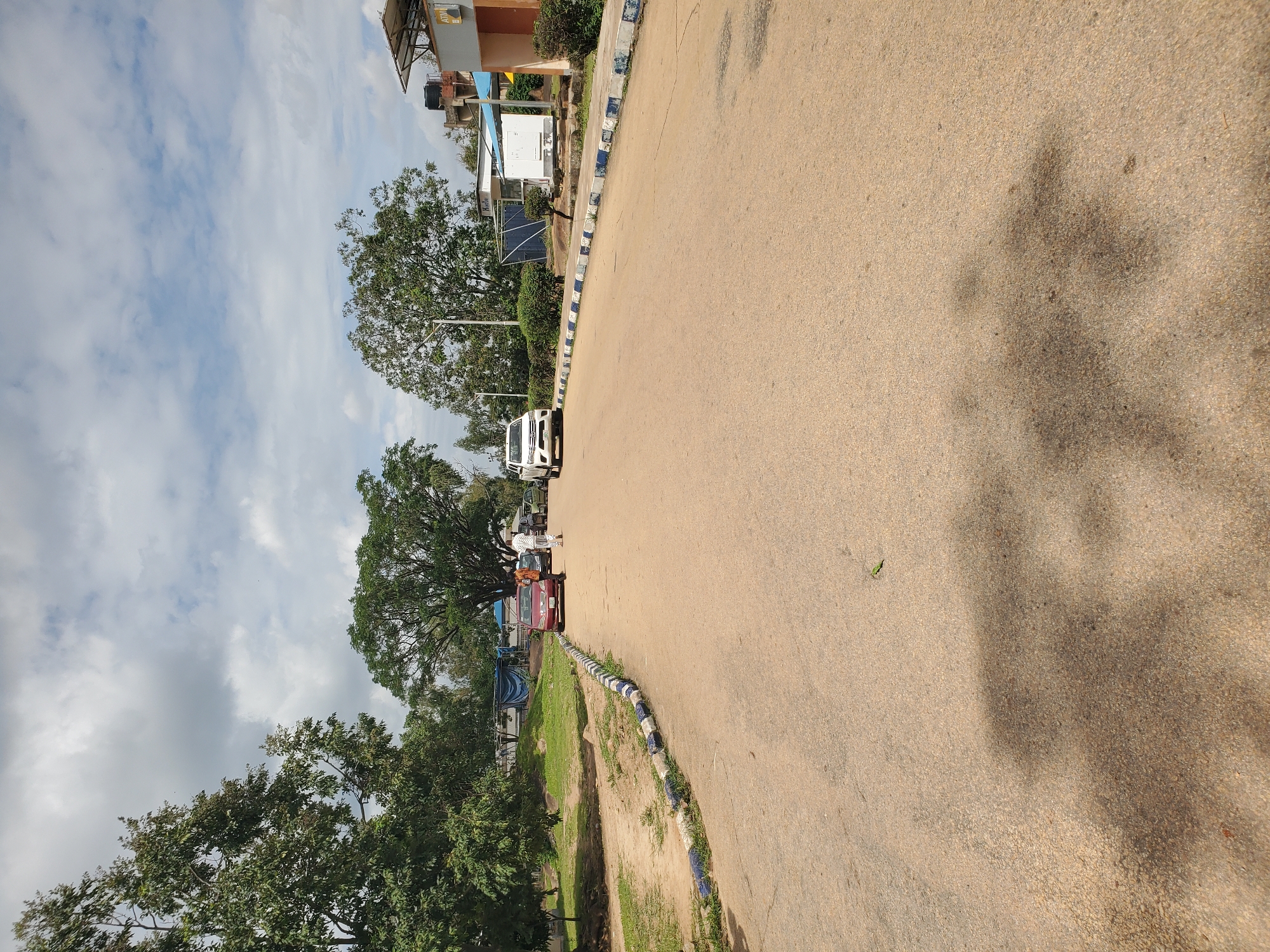 The image depicts an outdoor scene on a paved road in Jos, Nigeria, under a cloudy sky. The road, with a beige asphalt surface, is lined on the left and right with a curb decorated with blue and white painted stones. On the left side of the road, there is a grassy area with trees and some distant structures partially obscured by foliage. On the right side of the road, there are several buildings, including one with an ATM visible on its facade. A white SUV is parked on the right side of the road in the mid-ground, and a red sedan is parked on the left side, closer to the foreground. A man in a white robe and headscarf is standing between the two parked cars, and another person in an orange shirt is standing near the red car. Further down the road, there are more vehicles and people, suggesting a public or commercial area. Shadows of trees are cast across the road in the foreground.