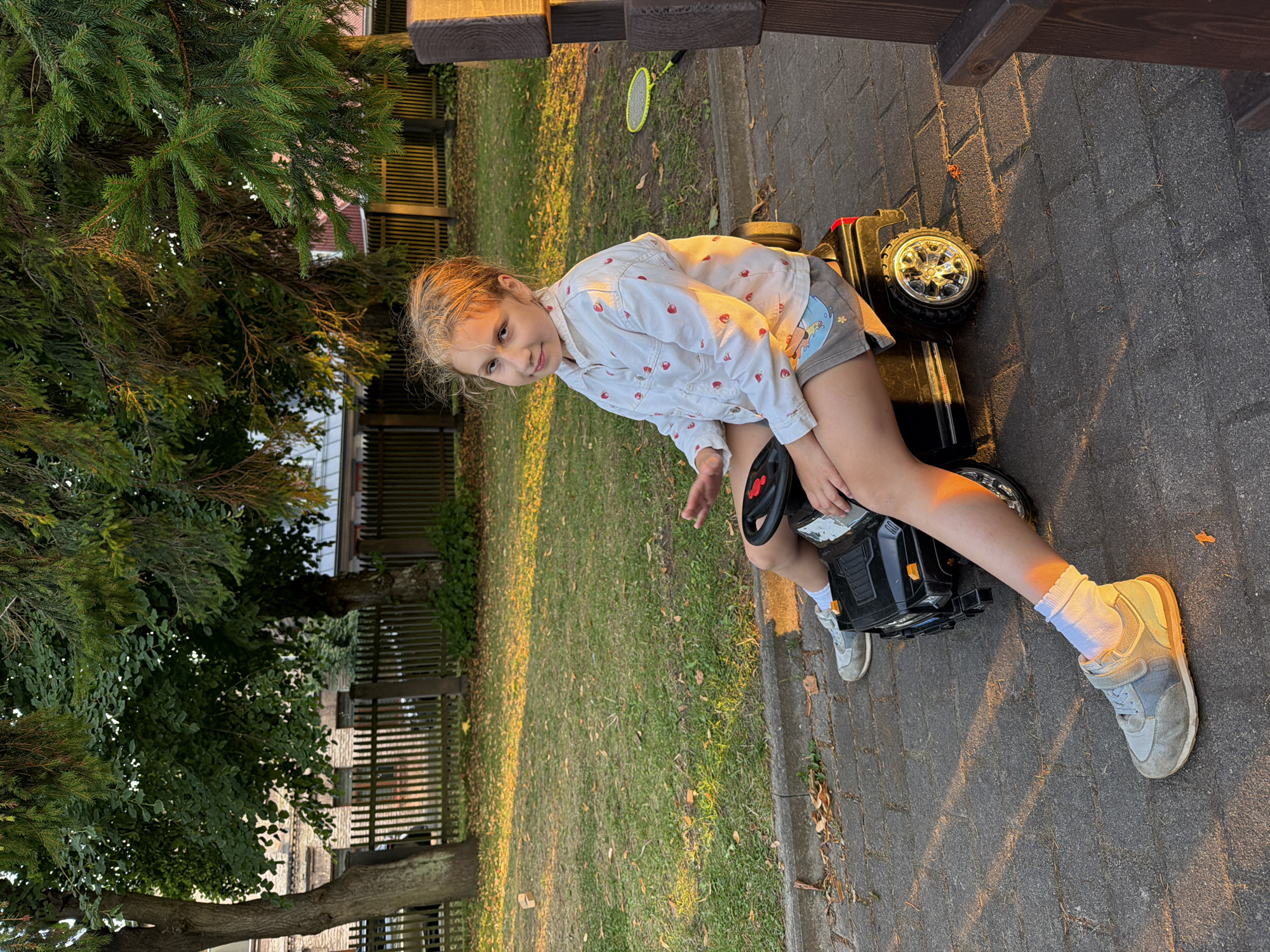 A young girl with blonde hair, wearing a white shirt with red strawberry patterns and grey shorts, sits on a black toy car. She is positioned on a paved area in front of a grassy lawn. Behind the lawn, a wooden fence separates the area from a street or pathway, with buildings visible beyond the fence. Trees, one with a thick trunk and green foliage, and another with pine needles, are prominent in the background, casting dappled sunlight across the scene. A badminton racket lies on the grass to the right of the toy car. The lighting suggests it is late afternoon or early evening, with long shadows cast by the trees. The location is the city of Ventspils, Latvia.