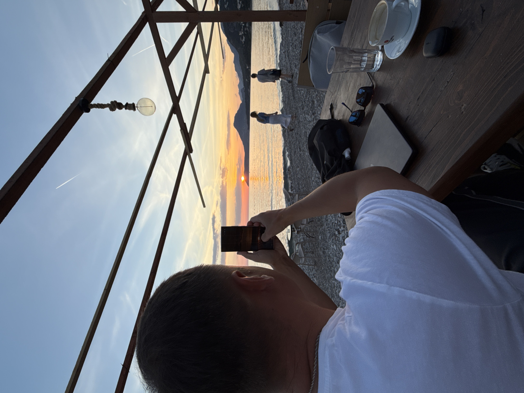 The image captures a sunset view over the sea from a partially covered outdoor seating area, likely in Bar, Montenegro, given the landscape. A person is taking a photo of the sunset with a smartphone. In the background, two people are walking along the shoreline. The foreground features a wooden table with a cup, a glass of water, sunglasses, a black pouch, and a black pad, suggesting a cafe or restaurant setting. The sky exhibits colors of orange and yellow. The seating area's structure is visible with wooden beams overhead.