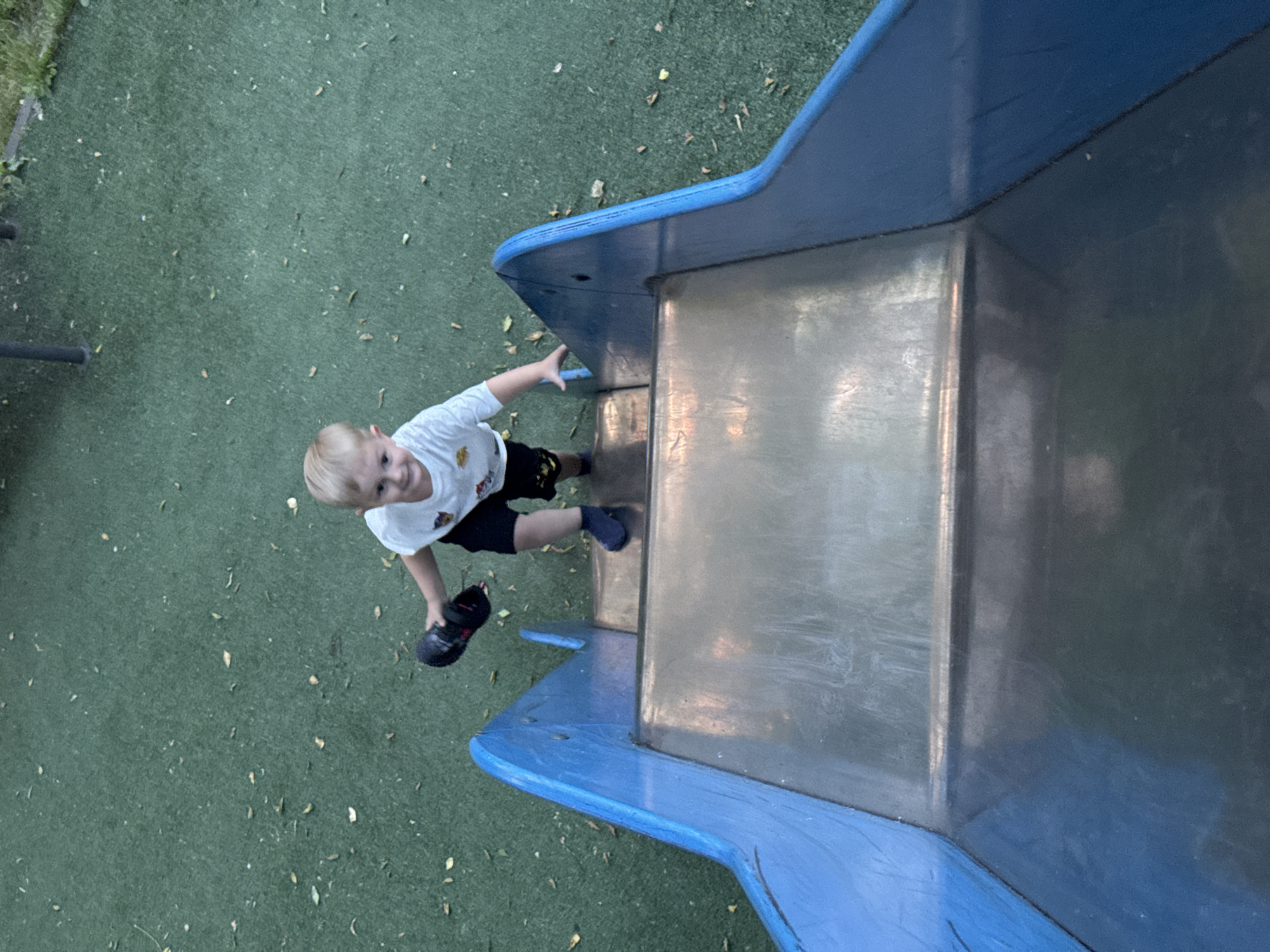 A young boy with blonde hair and a white t-shirt with a cartoon graphic is climbing up a blue metal slide. He is holding a black shoe in his right hand. The slide is on a green artificial turf surface, possibly in a playground or park. There are scattered leaves on the ground. The boy is looking towards the camera with a slight smile. The overall scene suggests a playful outdoor activity.