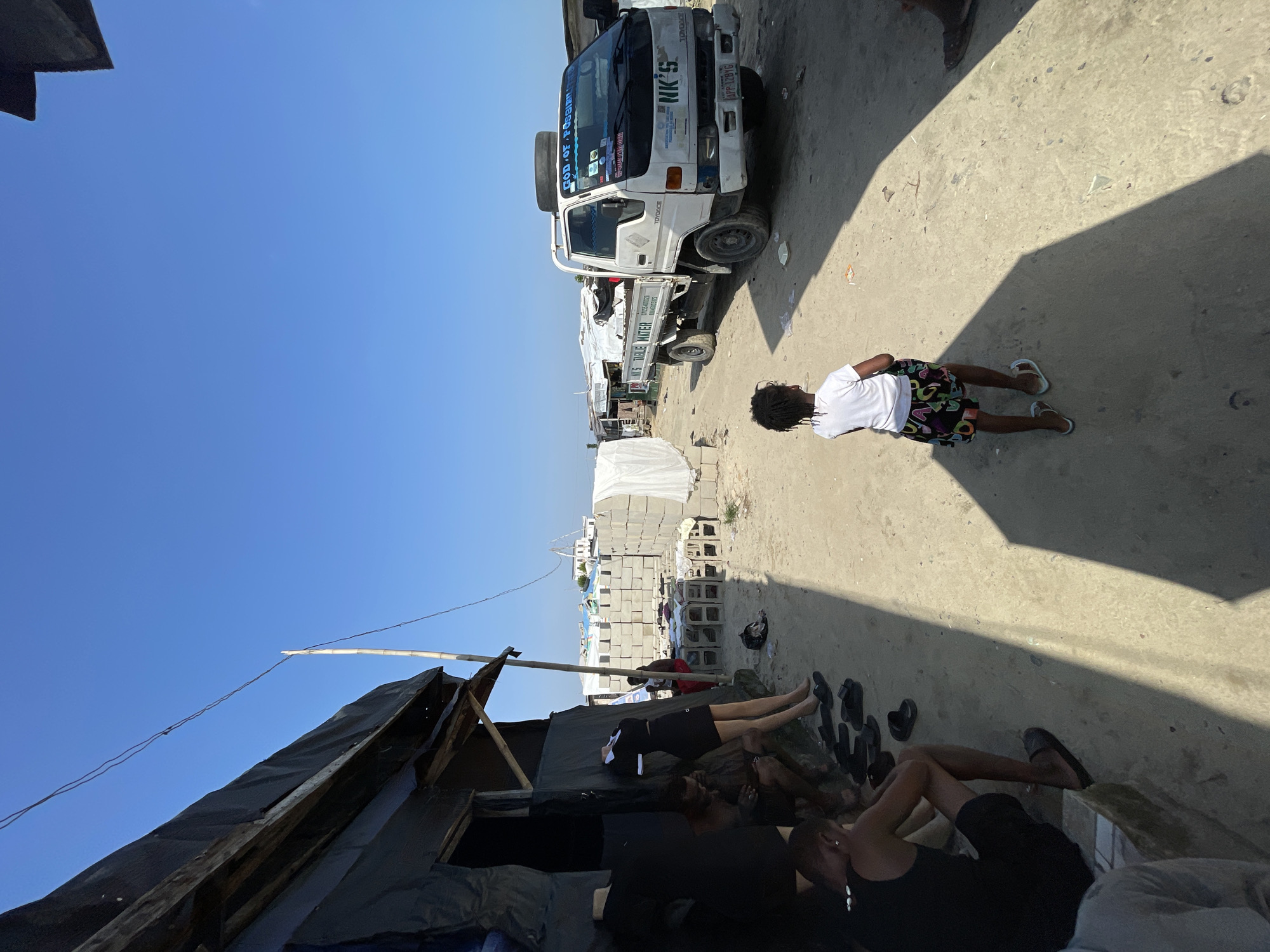In Iranla, Nigeria, a clear sky looms over a dusty, sun-drenched environment. A white flatbed truck, possibly a delivery vehicle, is parked on the right side of the frame. It displays the text "NK'S TABLE WATER" on its side and "NK'S" on the front. The truck is adorned with stickers, one of which reads "GOD OF POSSIBILITY." A young child, wearing a white t-shirt and a patterned skirt, stands in the foreground, facing away from the camera and towards the truck. To the left, a makeshift structure with a dark, tarp-like covering and exposed wooden beams suggests a humble dwelling or market stall. Several people are seated or standing near this structure, some appearing to be resting or engaged in conversation. In the background, stacks of concrete blocks and what appear to be additional structures indicate a developing or informal settlement. The ground is a mix of dirt and sand, with scattered debris. The overall scene depicts a snapshot of daily life in this Nigerian community.