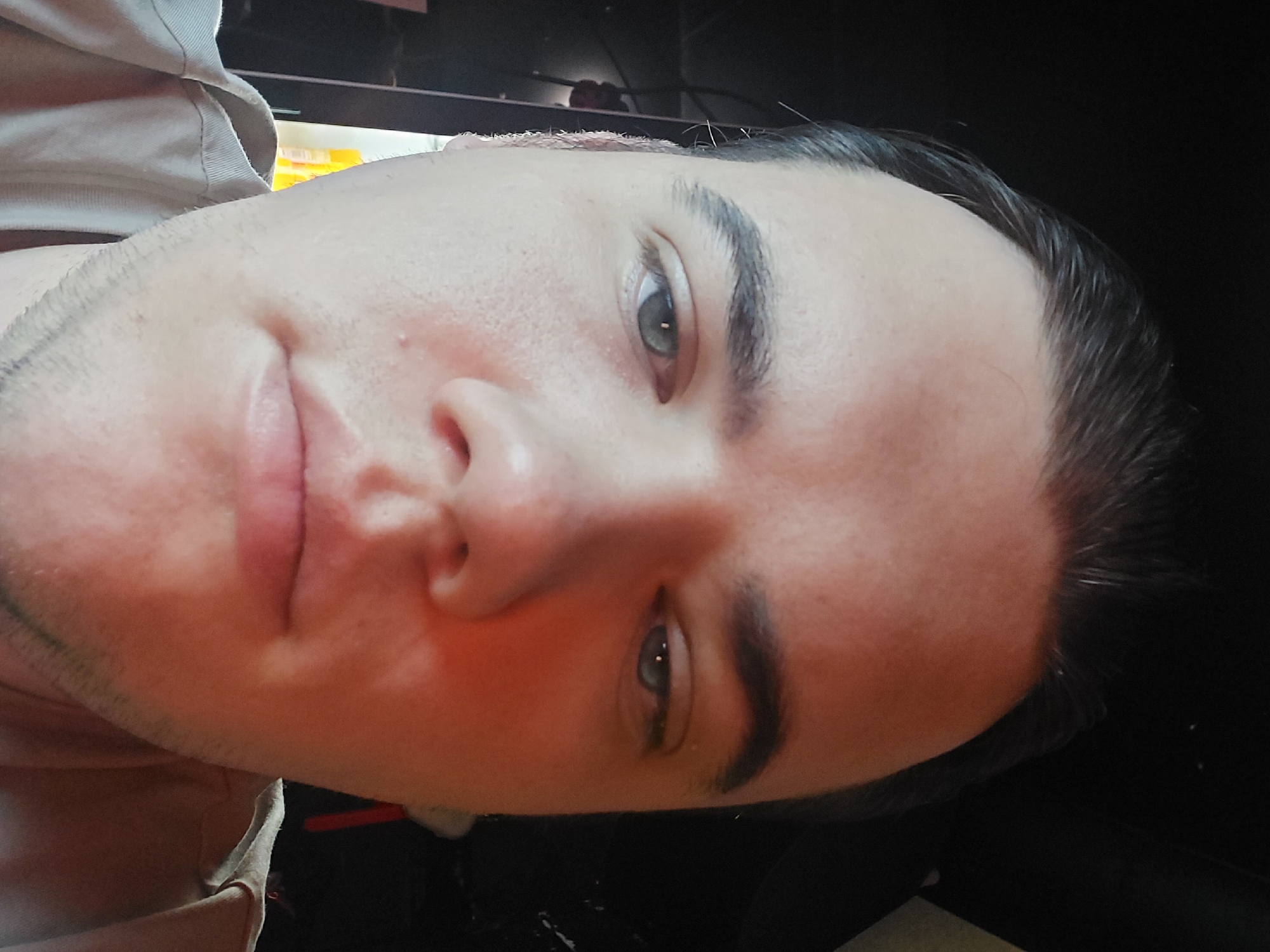 A close-up, slightly angled portrait of a fair-skinned individual with short, dark hair. The person has light-colored eyes and is giving a slight smile. The background is partially obscured, with a dark area to the right and what appears to be a blurred shelf with various objects to the left. Clothing is visible at the bottom left corner. There is no evidence to determine the location to be Tashkent, Uzbekistan.