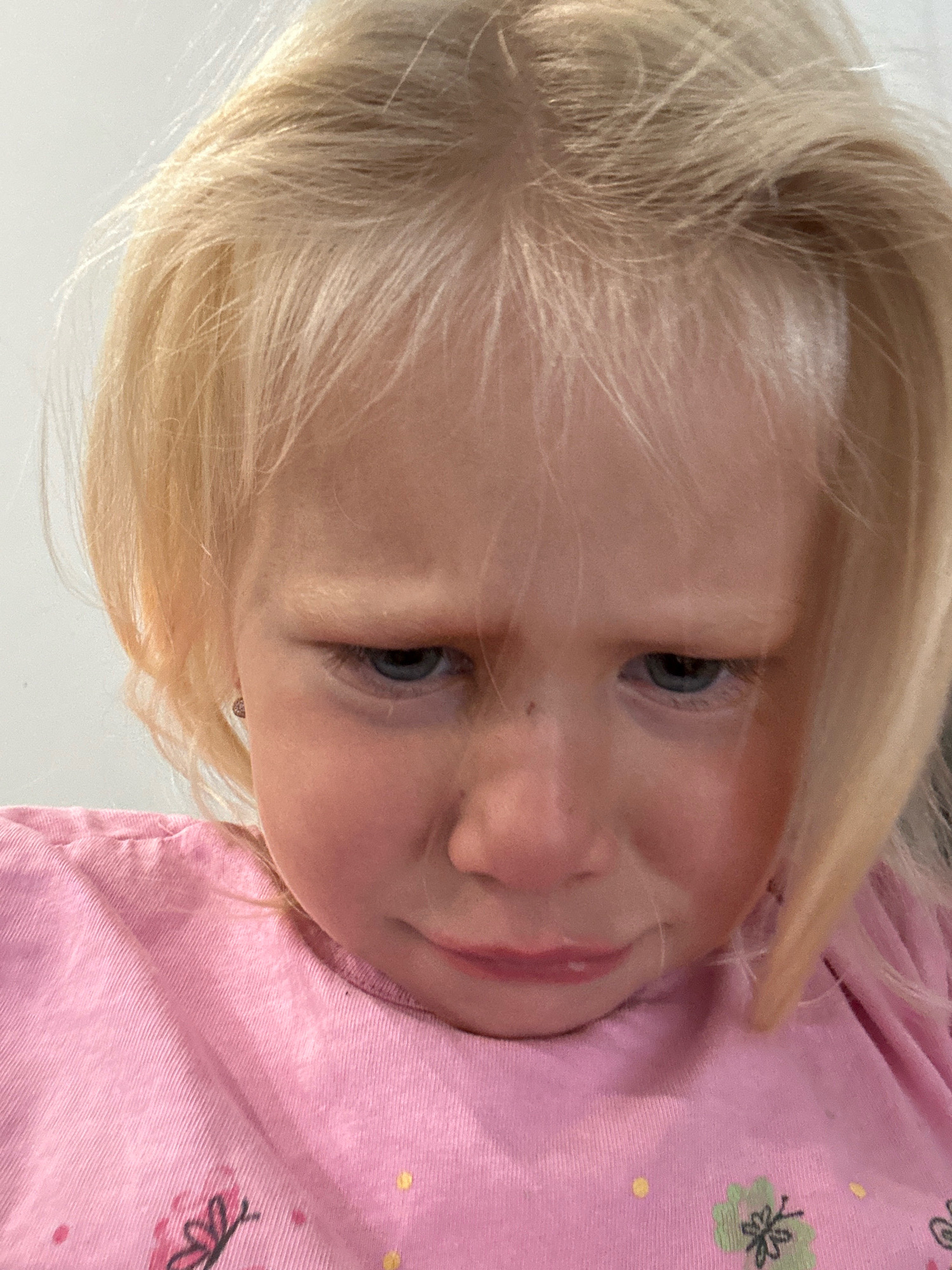 A close-up, overhead shot shows a young Caucasian girl with blonde hair and blue eyes. She is wearing a pink t-shirt with a floral design. Her expression is sad, with her eyebrows furrowed and her mouth slightly open. The background is a plain white wall. The image is taken indoors. There is no visible indication of the location being Milton Keynes, United Kingdom.