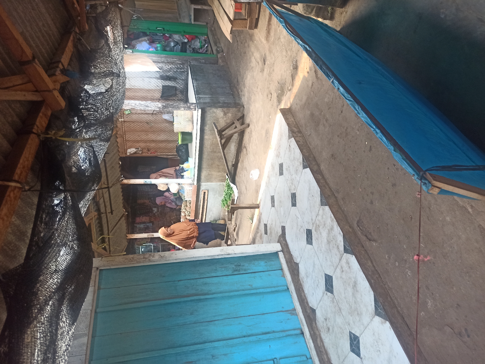 The image depicts an outdoor market stall area in Trisinar, Indonesia. Two women are visible at the stall. One woman, wearing a brown hijab and dark clothing, stands behind a counter, appearing to be tending to produce. Another woman, wearing a pink hijab, is partially visible further back at the stall. Various goods, including what appears to be pineapples and leafy green vegetables, are displayed on the counter and on a wooden bench in front of it.
The market stall has a weathered roof made of corrugated metal and wood, partially covered by black netting for shade. The ground is unpaved, with sandy dirt. To the left, a section of a blue wooden door or wall is visible, with a tiled walkway in front of it featuring a diamond pattern. In the foreground, a blue tarp is stretched out on a wooden frame, obscuring part of the ground. The overall environment suggests a simple, functional market setting, likely in a rural or semi-urban area of Indonesia.