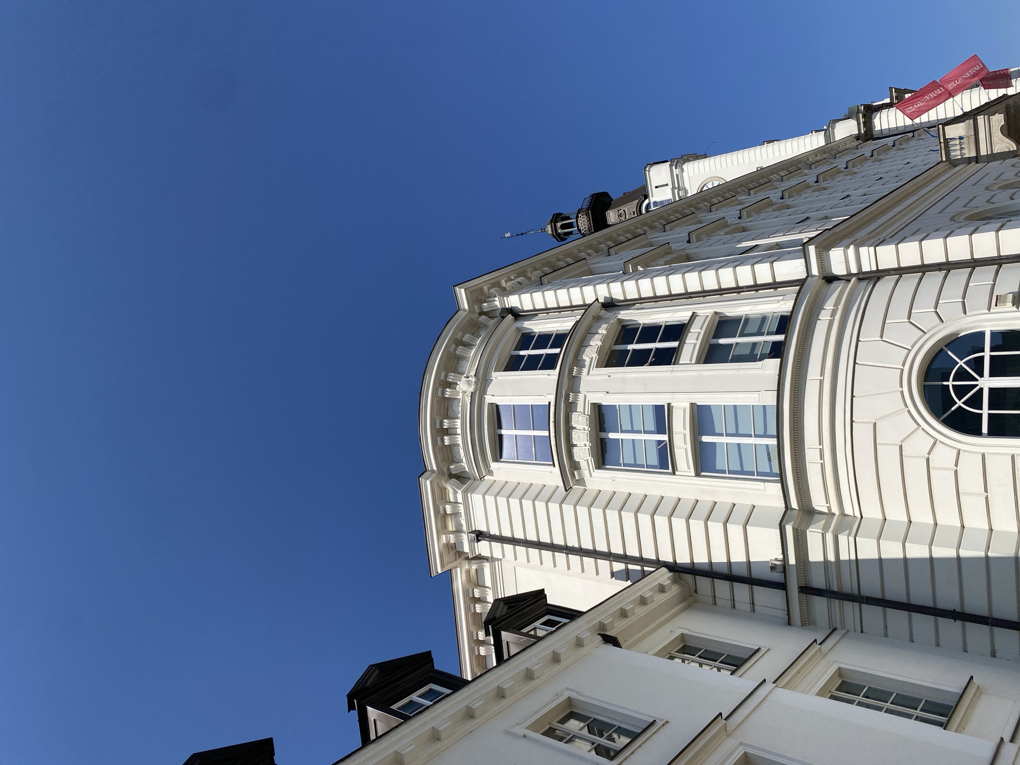 This is an exterior, low-angle, eye-level shot of a white, ornate building against a clear blue sky. The building has multiple windows with white frames and is adorned with detailed architectural elements. To the left, another white building is partially visible. A tower with a dark roof and spire is visible further down the street, along with two red banners that read "GENERALI". The scene is likely located in Warsaw, Poland, given the presence of the Generali building, a known landmark in the city. There are no people or animals visible in the image.