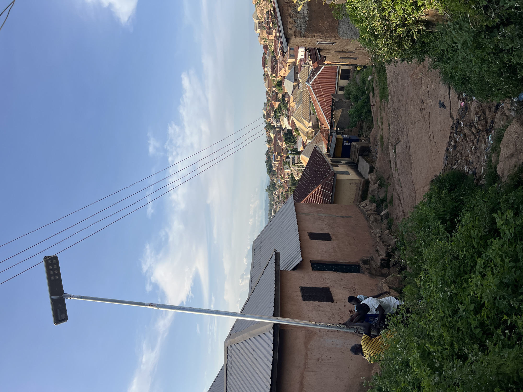 In Kabong, Nigeria, three young men are gathered around a tall pole, seemingly engaged in an installation or repair. The pole supports a solar-powered streetlight, its panel facing the sky. In the background, a densely populated hillside is dotted with houses, many featuring corrugated metal roofs. The uneven terrain is characterized by a dirt path winding downhill, bordered by lush green vegetation. The sky above is a vibrant blue, adorned with scattered white clouds.