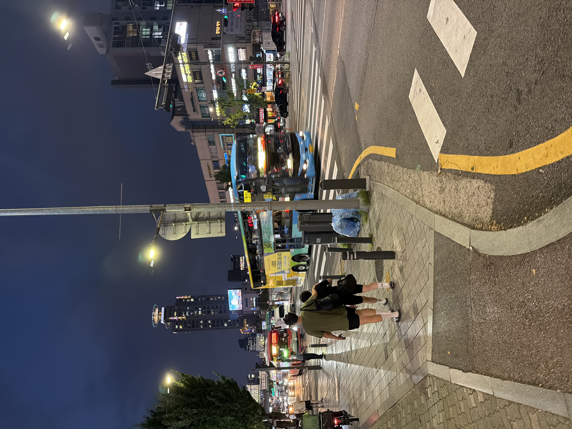 In this nighttime street scene in Seoul, South Korea, a double-decker blue bus moves across a pedestrian crossing, its headlights illuminating the wet asphalt. To the left, two figures, a man and a woman, walk away from the camera on a paved sidewalk, casting long shadows under the streetlights. Behind them, a red bus is stopped at an intersection, and other vehicles and buses are visible in the distance. Buildings line the street, with illuminated signs and windows indicating urban activity. A tall skyscraper with a helipad on top is prominently featured in the background, its lights piercing the dark sky. The overall environment suggests a busy city street at dusk or night, with traffic and pedestrian movement.