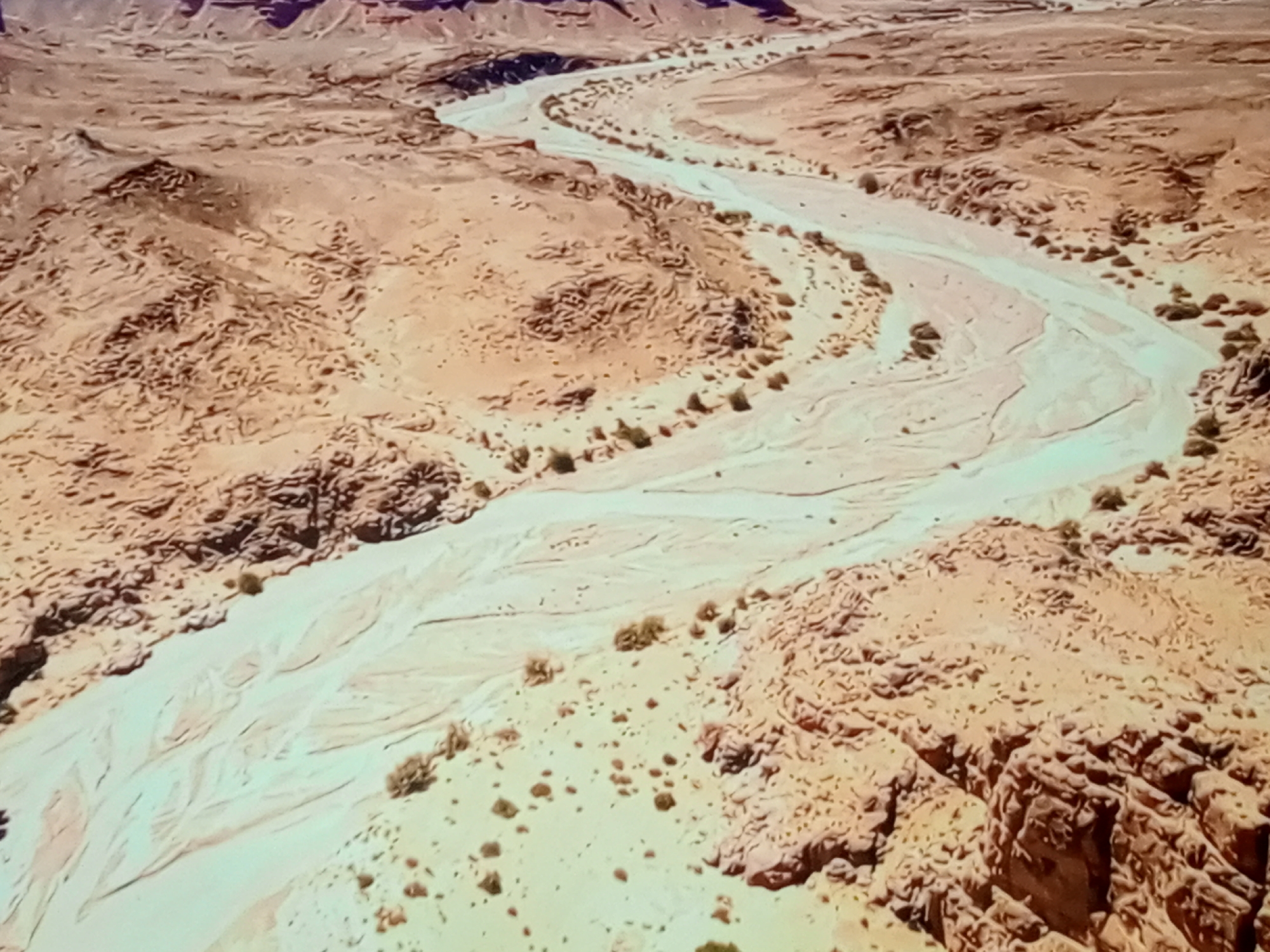 An aerial view shows a winding dry riverbed cutting through a desert landscape. The riverbed is a pale, sandy color, contrasting with the surrounding reddish-brown earth and rock formations. Sparse, low-lying vegetation is scattered throughout the scene. The terrain appears arid and rugged, with layered rock formations visible in the distance. The overall impression is one of vast, natural beauty in a harsh environment.
