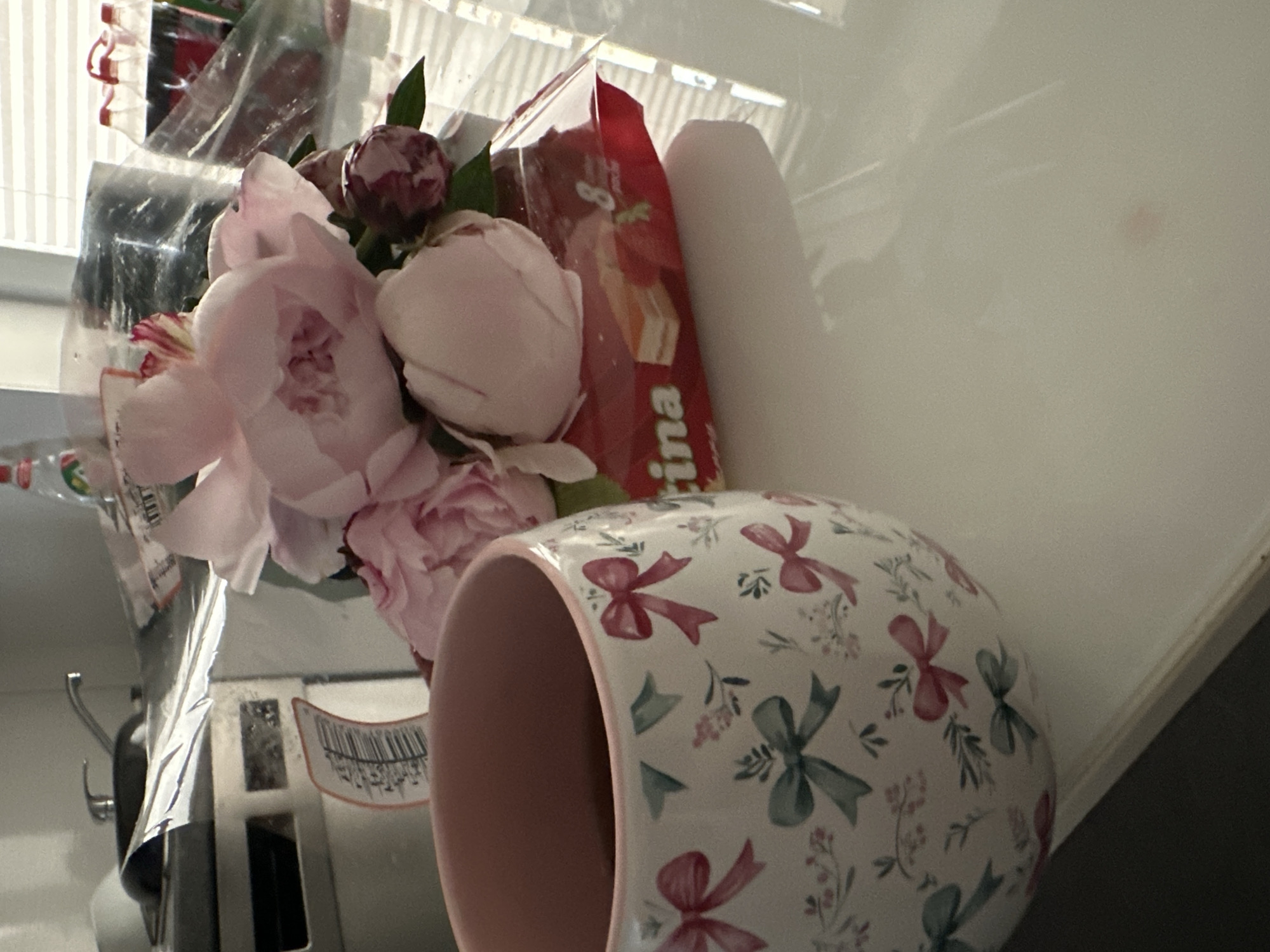 The image depicts a close-up of a table with a coffee mug adorned with a floral and bow pattern. Next to the mug are several pink peonies and a package of biscuits, partially wrapped in plastic. In the background, a kitchen sink and faucet are visible, along with plastic-wrapped bottles of soda. The scene appears to be set indoors, possibly in a kitchen or dining area. No specific location in Kutenholz, Germany is identifiable from the image itself.