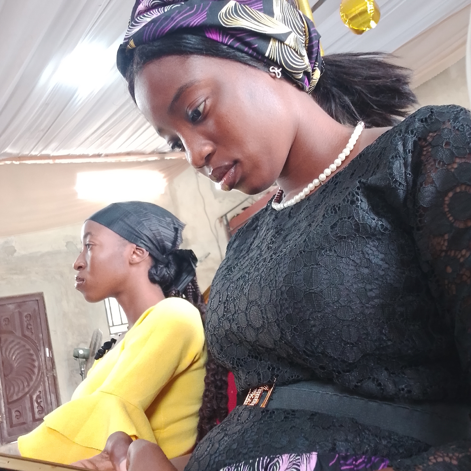 The image shows two young women from the chest up, seated indoors in what appears to be a church hall in Ado, Nigeria. The woman in the foreground on the right is looking down and to her left, wearing a black lace dress, a pearl necklace, and a purple and yellow patterned headscarf. She has dark skin and dark hair tied back. The woman on the left is looking straight ahead, wearing a bright yellow blouse with ruffled sleeves and a black headscarf. She also has dark skin and dark braided hair. The background shows a white ceiling, a closed wooden door with ornate carvings on the left, a wall with a textured finish, and a fan in the middle ground. A gold balloon is visible in the upper right corner of the image. The lighting suggests an indoor setting, possibly with natural light coming from an unseen source.
