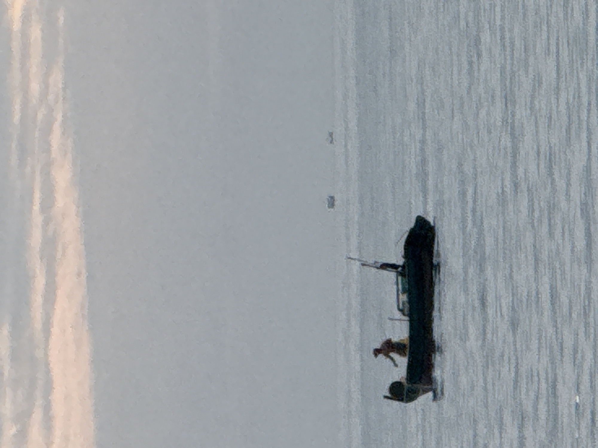 The image depicts a serene seascape with a soft, overcast sky, tinged with pale pink and white clouds. The sea is calm, reflecting the muted tones of the sky. In the foreground, a dark-hulled boat is prominently featured, with a lone figure, presumably a fisherman, standing within it. The fisherman is wearing a hat and appears to be engaged in an activity related to fishing. The boat has a dark tarp covering part of its interior and features a mast or pole towards the stern. In the distance, several other smaller boats are visible on the horizon, appearing as faint silhouettes against the hazy sea and sky. The overall atmosphere is tranquil, suggesting early morning or late evening light, typical of coastal areas like Nha Trang, Vietnam, where such scenes are common.