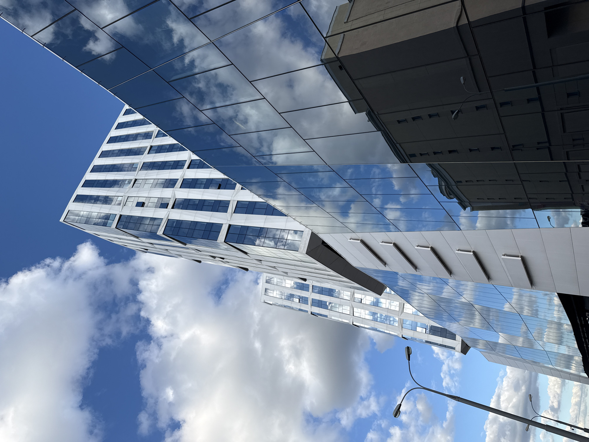 The image shows a low-angle view of modern architecture under a blue sky with scattered clouds. A tall, white building with a grid of windows dominates the left side of the frame, its design oriented upwards. Above and to the right, a glass structure reflects the sky and nearby buildings, creating a mirrored effect. The structures, situated in the city of Moscow, Russia, feature reflective properties, mirroring the sky and surroundings. At the bottom right, a gray lamppost extends partially into the frame.