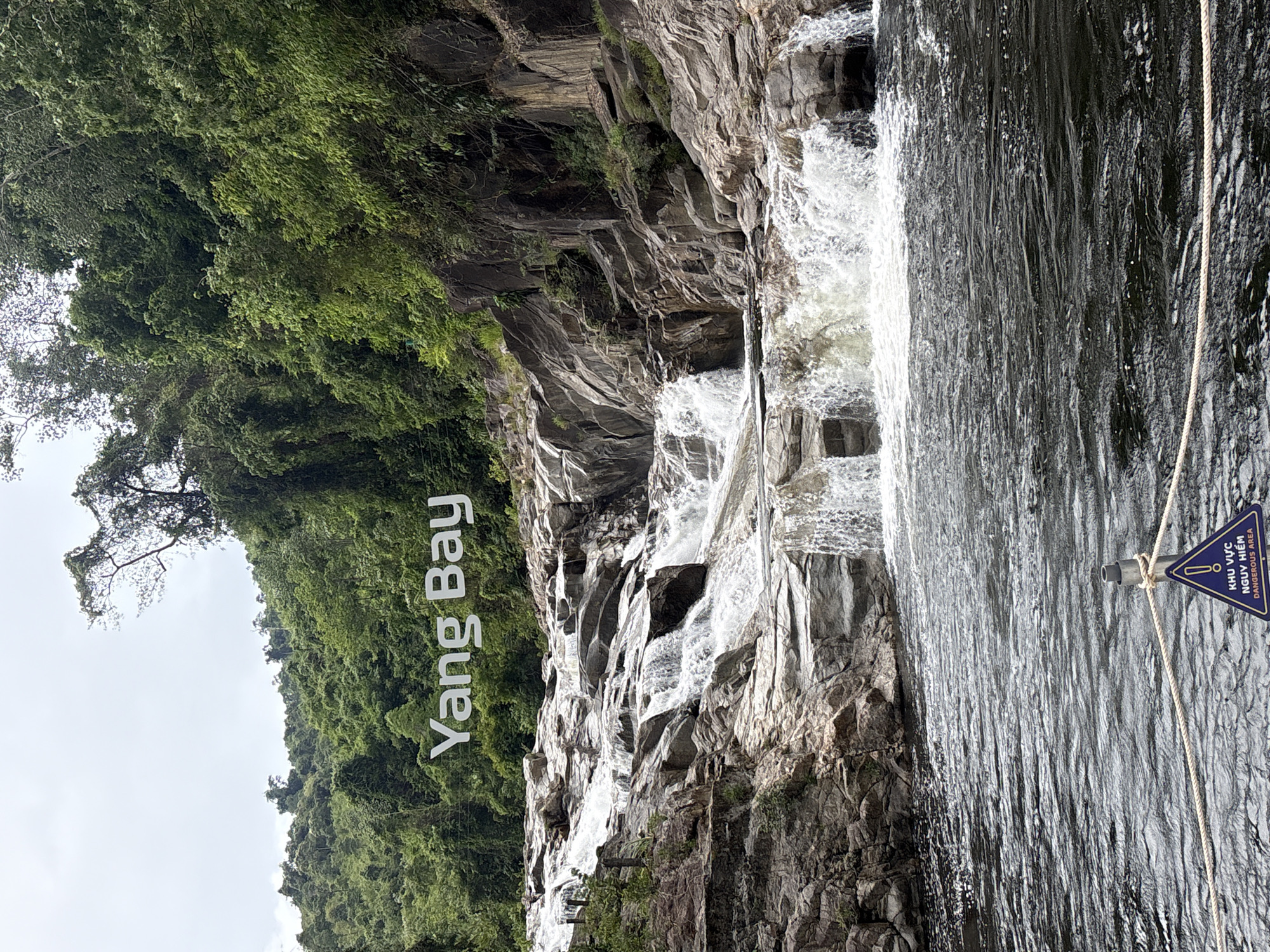 A wide shot captures the "Yang Bay" sign in large white lettering on a forested mountainside above a cascading waterfall in Khanh Vinh, Vietnam. The foreground features dark, rippling water. A triangular warning sign with a symbol and text reading "KHU VUC NGUY HIEM DANGEROUS AREA" is partially visible in the lower right corner, attached to a rope barrier. The waterfall flows over tiered, grey rocks, with lush green trees and foliage covering the surrounding hills. The sky is overcast.