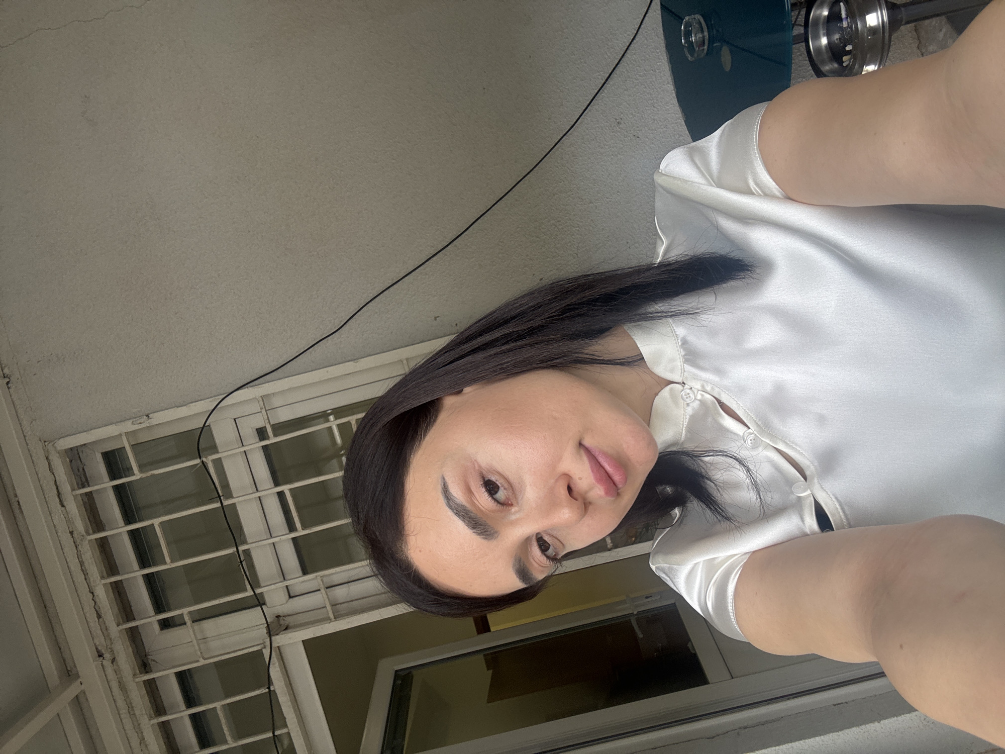 A selfie shows a woman with dark hair wearing a white satin top. She is indoors, near a window with a metal security grill, likely in Tbilisi, Georgia, judging by the architectural style and window construction. The background suggests an urban setting. A portion of a green object with metallic elements is visible in the upper right corner, potentially a piece of furniture or equipment.
