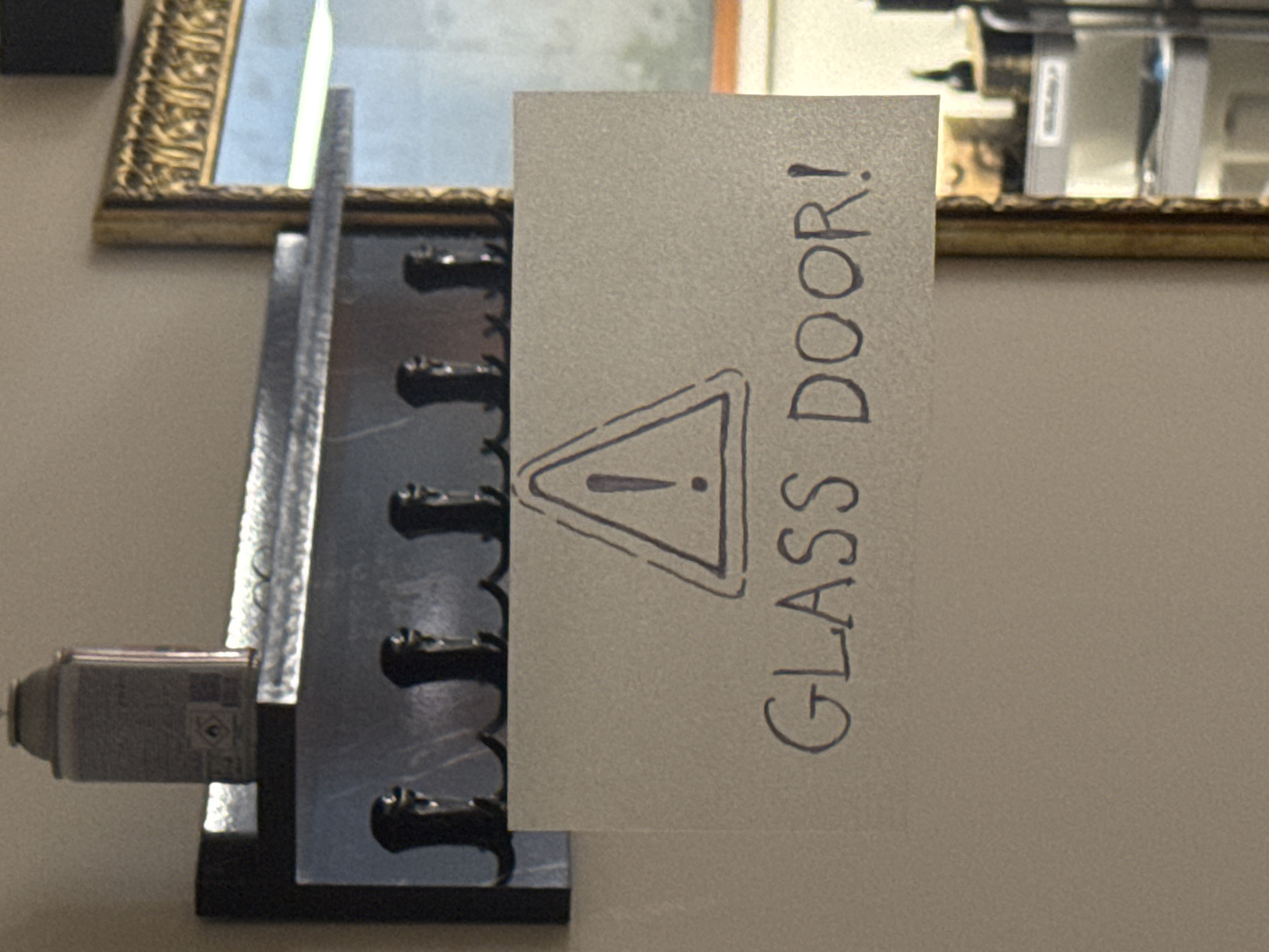 A close-up shot shows a sign on a wall that reads "GLASS DOOR!" with a hand-drawn warning triangle containing an exclamation point. The sign is attached to a coat rack with multiple hooks. In the background, a gilded mirror reflects a room with shelves and equipment. The image appears to be taken indoors, possibly in a commercial establishment. The specific location in Sofia, Bulgaria, is not identifiable from the visual information provided.