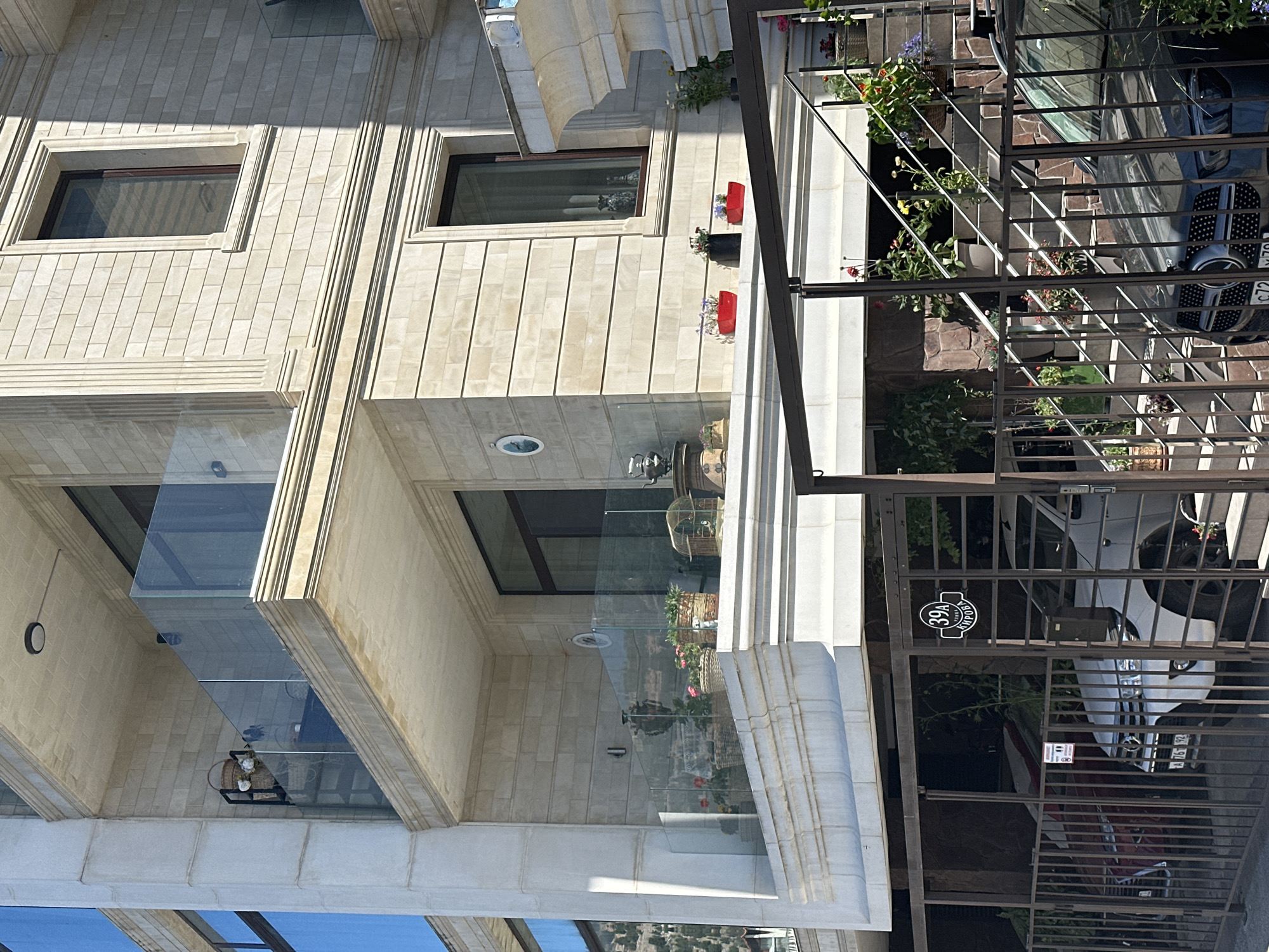 The image shows a multi-story building with a facade of light-colored stone blocks. The building has several balconies with glass railings. There are potted plants on the balconies and near the entrance. A staircase with metal railings leads up to the building. In the foreground, there is a metal fence and parked cars, including a white SUV and a dark sedan. A street sign with the number "39A" and some text is visible on the fence. The environment appears to be an urban or suburban setting.