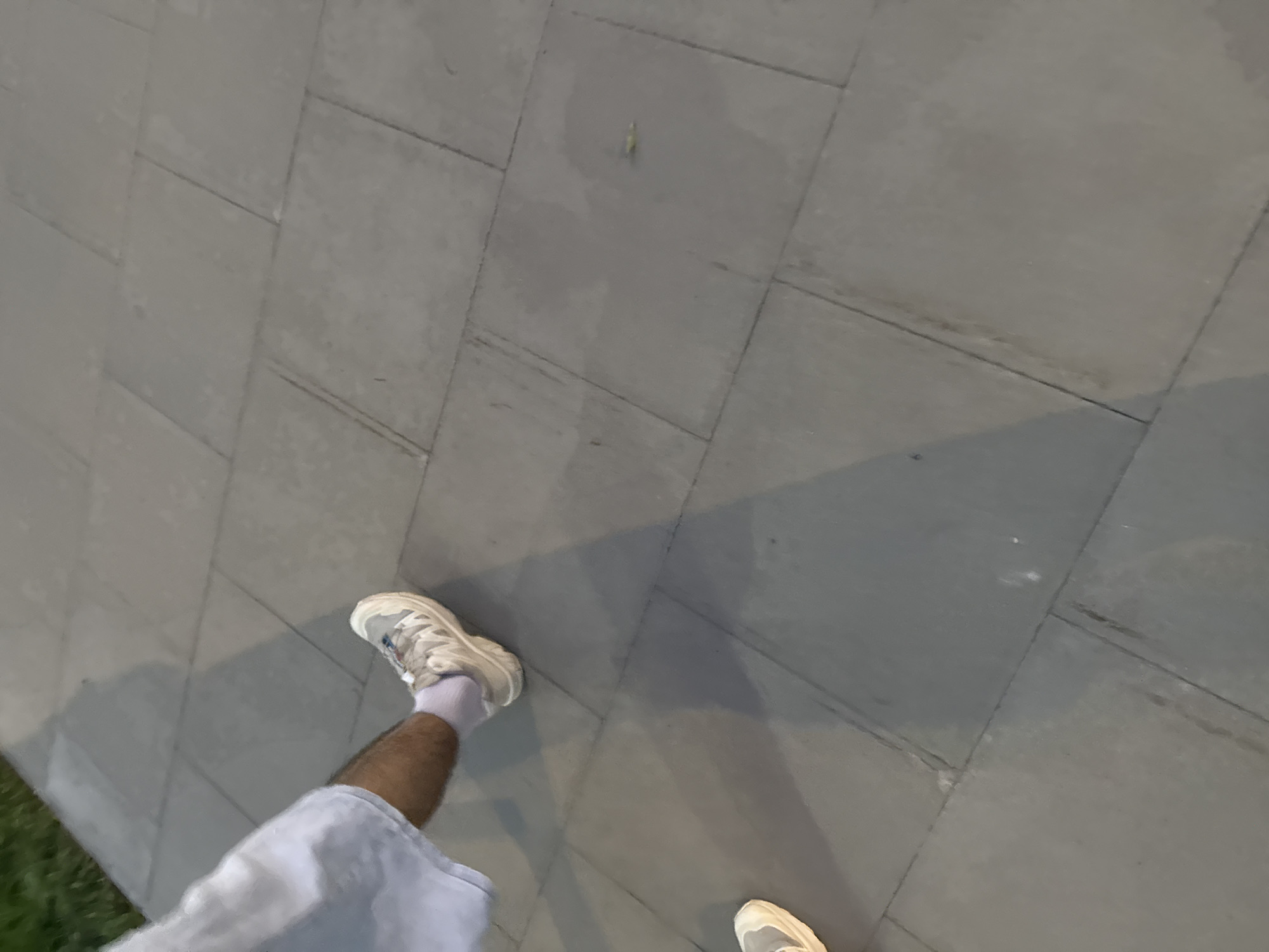 A person is walking on a paved walkway. The walkway is made of large, square, grey stone tiles. The person is wearing light blue shorts, white socks, and light-colored athletic shoes. Their right leg is extended forward, and their left foot is positioned behind it. The ground casts a shadow on the walkway, indicating an external light source. To the left of the walkway, there is a patch of green grass.