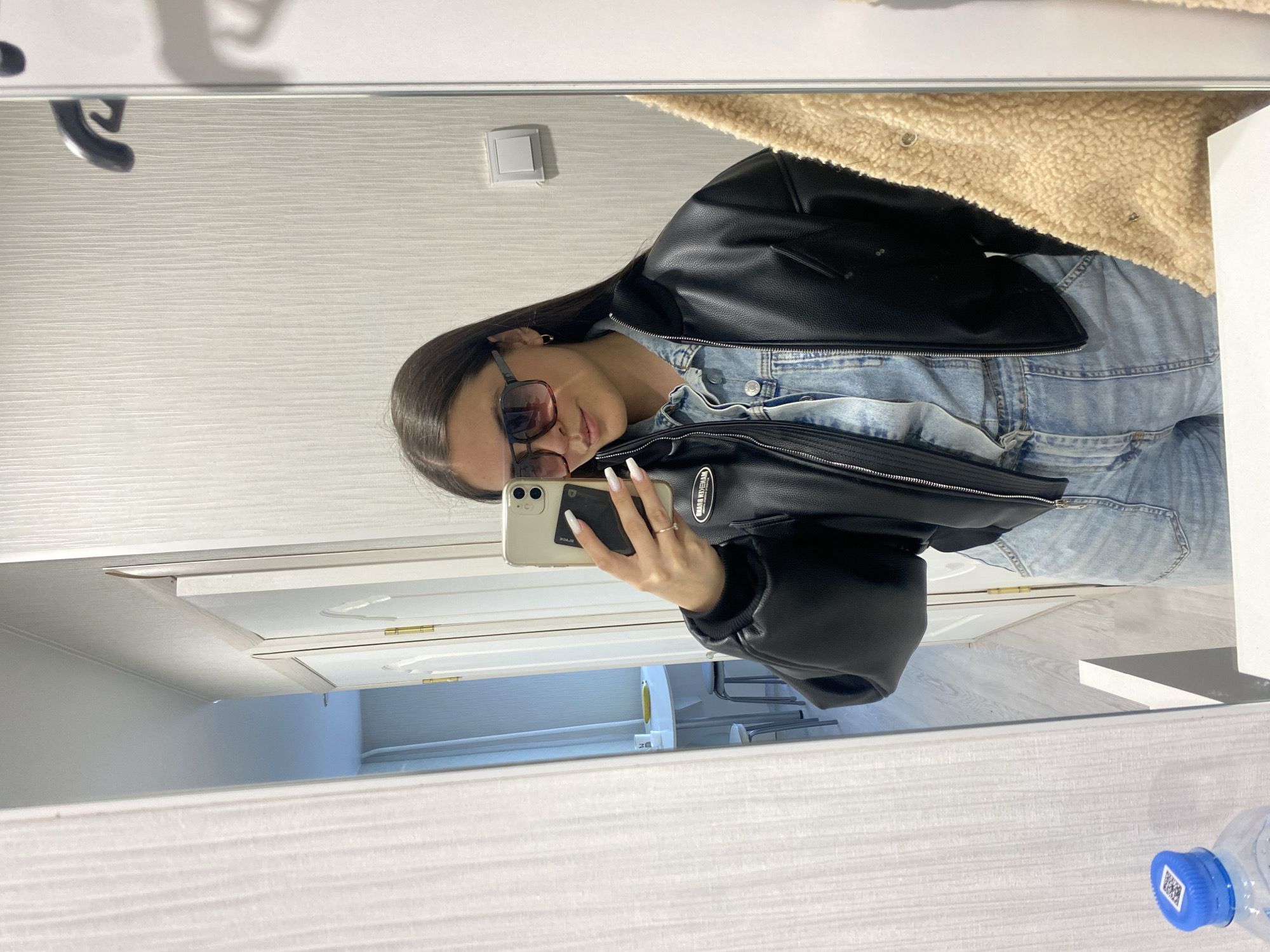 A selfie photograph shows a woman in a mirror's reflection. She wears sunglasses, a denim jumpsuit, and a black leather jacket. The environment includes a white wall, an electrical outlet, and a door. The location is suggested to be Chaikovsky, Russia, based on the presence of Cyrillic script on an object visible in the mirror's reflection. A plastic water bottle sits on a white surface.