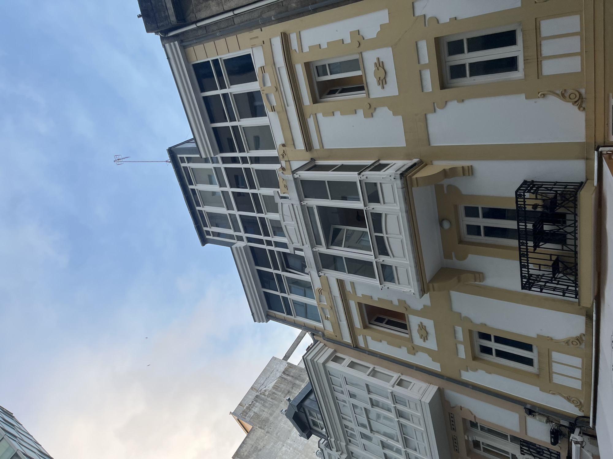 The image shows a low-angle view of a multi-story building with a distinctive architectural style, likely located in A Coruña, Spain. The building is predominantly white with yellow or light-brown accents highlighting its architectural details. Notable features include protruding glass-enclosed balconies on the upper floors, gridded windows, and decorative molding. A small black wrought-iron balcony is also visible. An antenna is attached to the top of the building. Part of a neighboring building and the blue sky with some white clouds is also visible.