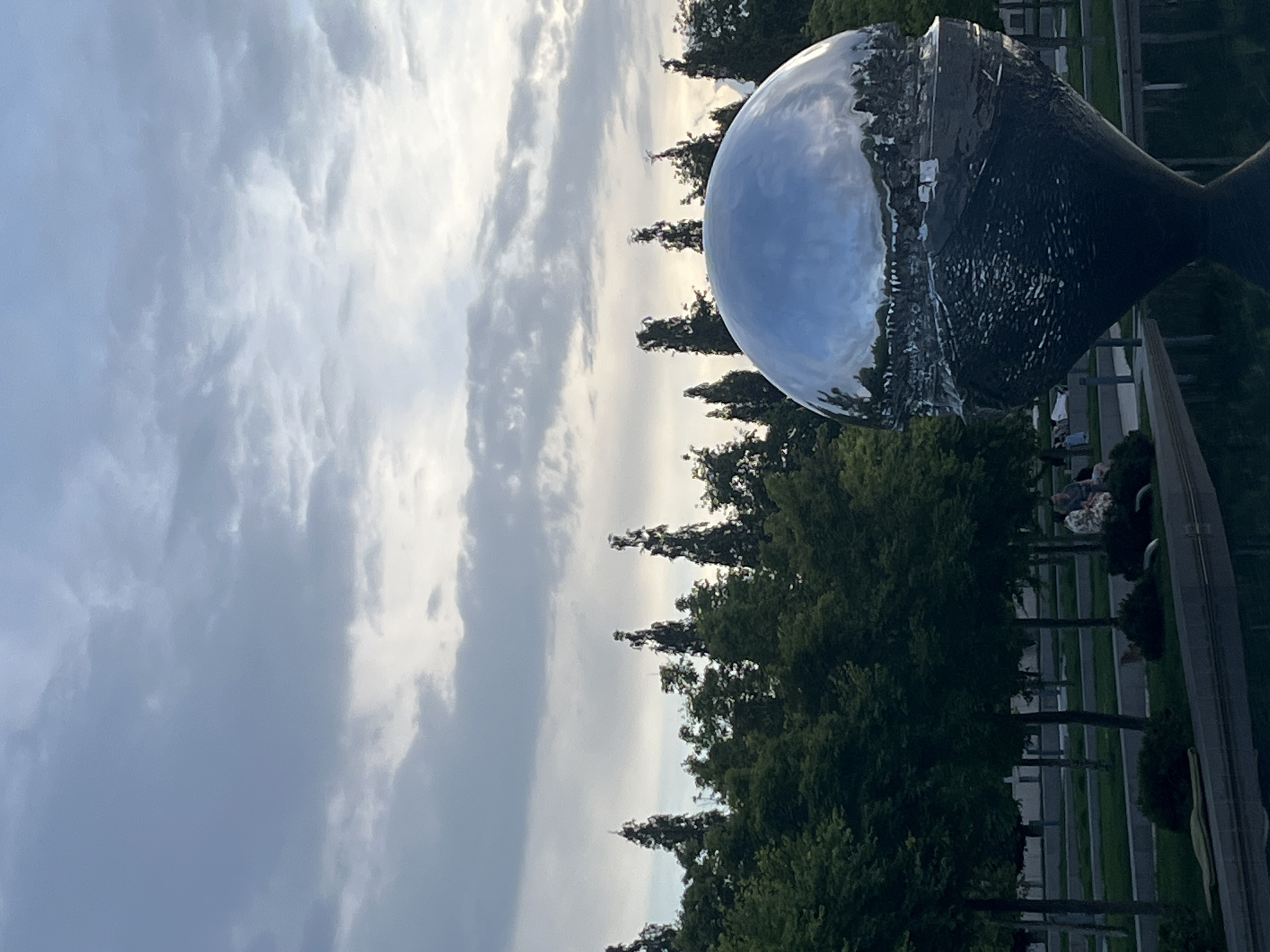 The image depicts an outdoor scene in Krasnodar, Russia, featuring a large, mirrored, spherical sculpture. The sculpture reflects the surrounding environment, including the sky with scattered clouds and nearby trees. The sculpture rests on a dark, metallic base, which appears to be a cone shape. In the background, there is a row of trees lining a walkway, a road, and some indistinct figures of people. The light suggests it is daytime.