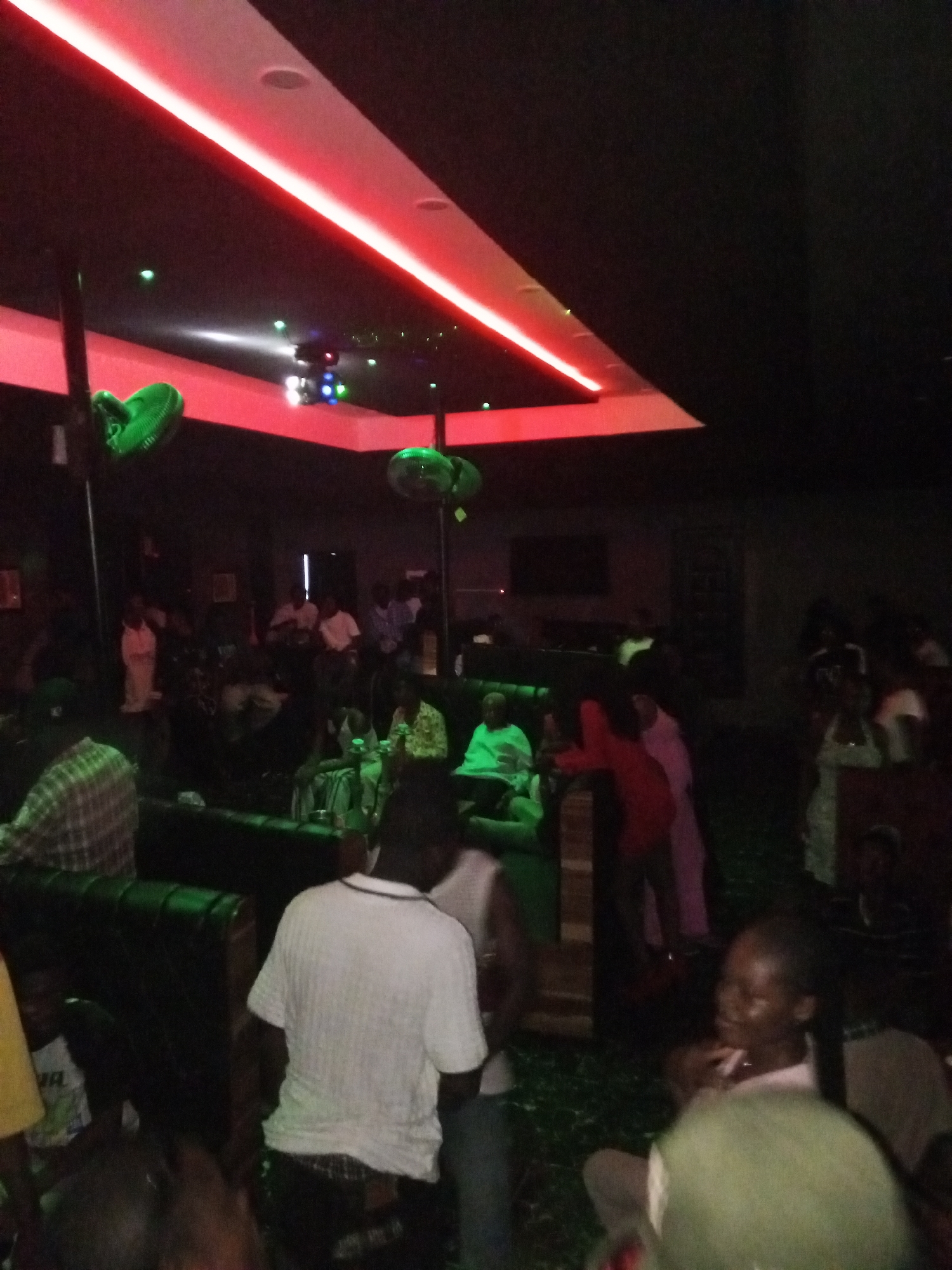 A dimly lit interior view shows a crowded space with multiple people, likely a club or lounge in Lokoja, Nigeria, given the context. The room has dark furnishings, including seating areas. The ceiling features a recessed section with red lighting, complemented by green decorative disco balls. A disco ball and other light fixtures are present. Most of the people are not clearly visible due to the low lighting and obstructions, but they appear to be engaged in conversations and general activity typical of such a venue.