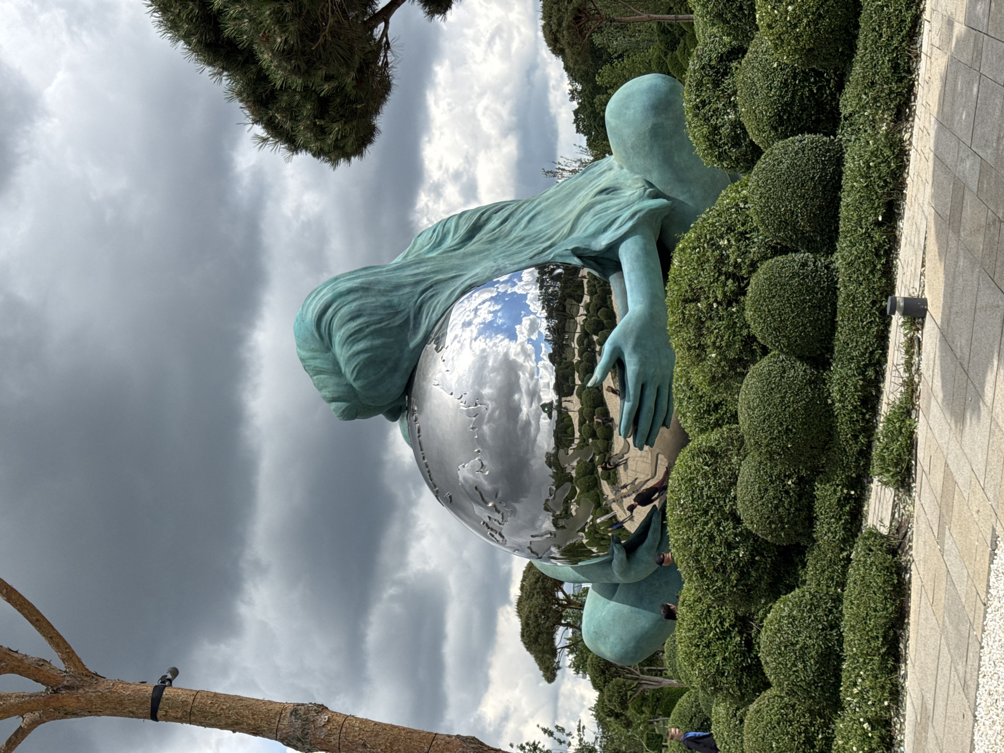 A vertically oriented photograph captures a statue of a kneeling female figure holding a spherical globe in a city park in Krasnodar, Russia. The statue is positioned amongst meticulously trimmed, rounded bushes. The sculpture is a teal color, and the globe has a mirrored surface. The sky above is filled with heavy, gray clouds. The reflection in the globe reveals the environment, including trees and people. A tree branch with a black strap is visible in the lower left corner of the frame, and a gray brick walkway lines the right side of the bushes and sculpture. The statue dominates the center of the image, with the cloudy sky in the background.