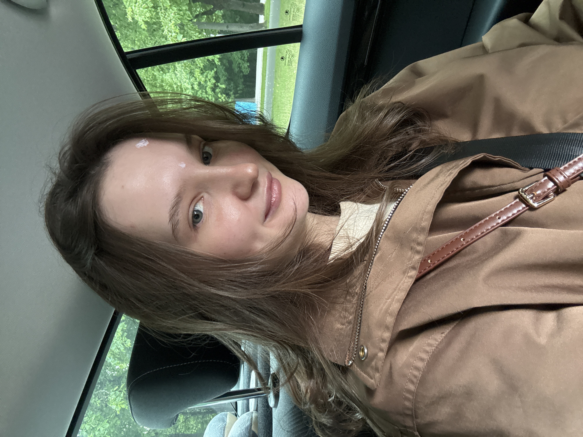 A young woman with long brown hair and fair skin is seated in a car, presumably the passenger seat, given the angle of the seat and the window. She is wearing a brown jacket and a visible seatbelt. There appears to be some cream or lotion on her forehead. The car is moving, as evidenced by the blurred greenery visible through the window. No specific information suggests this location is in Moscow, Russia.