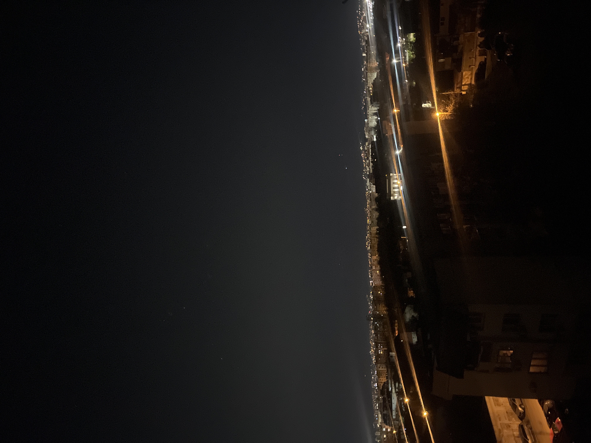 This is a night cityscape view of Sofia, Bulgaria, from an elevated position. The right side of the image shows buildings, trees, and streets with streetlights and car lights. The left side of the image is dark, likely depicting the sky or a large open space.