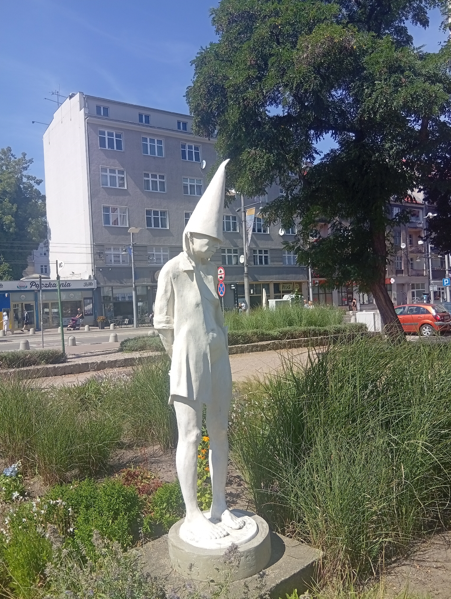 The image shows a white statue of a gnome with a tall, pointed hat standing in a garden. The statue is in the foreground, and behind it is a street with buildings and trees. The buildings are modern, with multiple stories and many windows. One building has the words "Poczta Polska" (Polish Post) and "Piekarnia" (Bakery) visible on its facade. An orange car is parked on the street. The statue is in Gdynia, Poland.