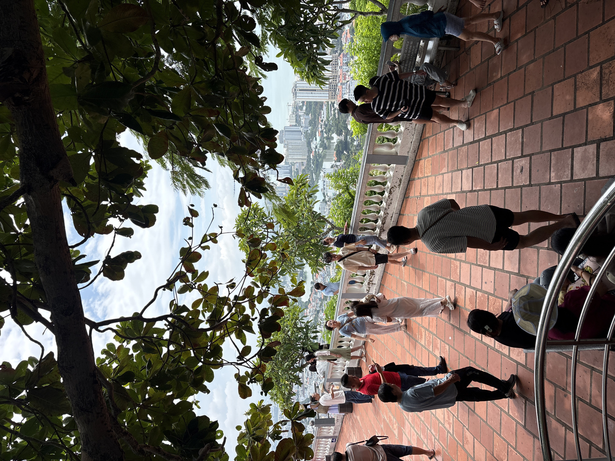 The image depicts a group of people on a tiled terrace overlooking a coastal city. The terrace is paved with reddish-brown tiles arranged in a diagonal pattern. A white balustrade runs along the edge of the terrace, providing a railing for visitors. Beyond the balustrade, a dense urban landscape unfolds, characterized by numerous buildings of varying heights, including high-rise structures and residential blocks. A body of water, likely a bay or inlet, is visible within the city, with boats dotting its surface. Lush green trees with large leaves overhang the terrace from above, creating a canopy that partially obscures the sky and the city view. The sky is partly cloudy.
Several individuals are present on the terrace, engaged in various activities. Some are standing and conversing, others are walking, and a few appear to be taking in the view. One person in the foreground is seated. The people are dressed in casual attire, suggesting a tourist or recreational setting. The overall scene conveys a sense of a popular vantage point, offering panoramic views of the city and its waterfront. The architecture of the balustrade and the tiled flooring suggest a cultural or historical site, potentially a temple, pagoda, or monument, given the context of Ho Chi Minh City, Vietnam.