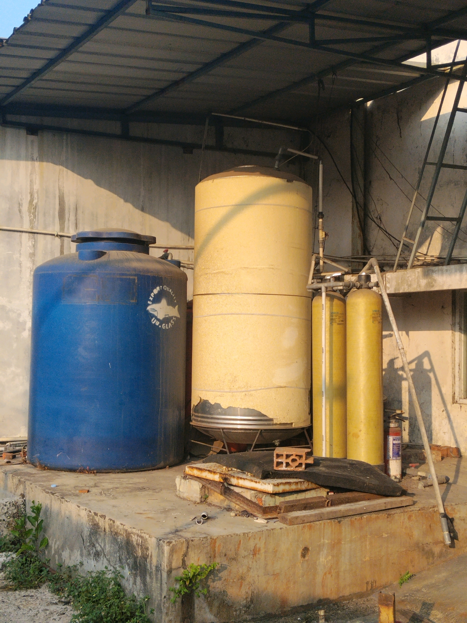 The image depicts an outdoor storage area, likely for water tanks, situated in Suka Nagara, Indonesia. The scene is dominated by two large industrial water tanks: one is a prominent blue cylindrical tank with a domed top and a circular emblem featuring a shark and the text "EXPORT QUALITY UB. GLASS" on its side. Adjacent to it is a taller, pale yellow cylindrical tank, also with a domed top, showing signs of wear and tear, including peeling paint or material near its base.
In front of these tanks, and to the right, are two upright yellow cylindrical vessels, likely part of a filtration or purification system. These are connected by a network of pipes and valves. A red and white fire extinguisher stands vertically near the yellow cylinders, leaning against the wall.
The tanks are positioned on a raised, weathered concrete platform. Scattered on and around the platform are various discarded items, including metal scraps, wooden planks, and a red brick. Green vegetation, consisting of small plants and weeds, grows around the base of the concrete structure and on the left side of the image.
Above the tanks and platform, a corrugated metal roof is supported by a metal frame, providing partial shelter. The background consists of a textured concrete wall, a metal ladder leaning against it on the right, and electrical wires strung across the area. The lighting suggests it is late afternoon or early morning, with long shadows cast by the objects and the structures. The overall impression is one of a functional, possibly industrial or agricultural, storage and processing area that has seen some use and may be somewhat neglected.